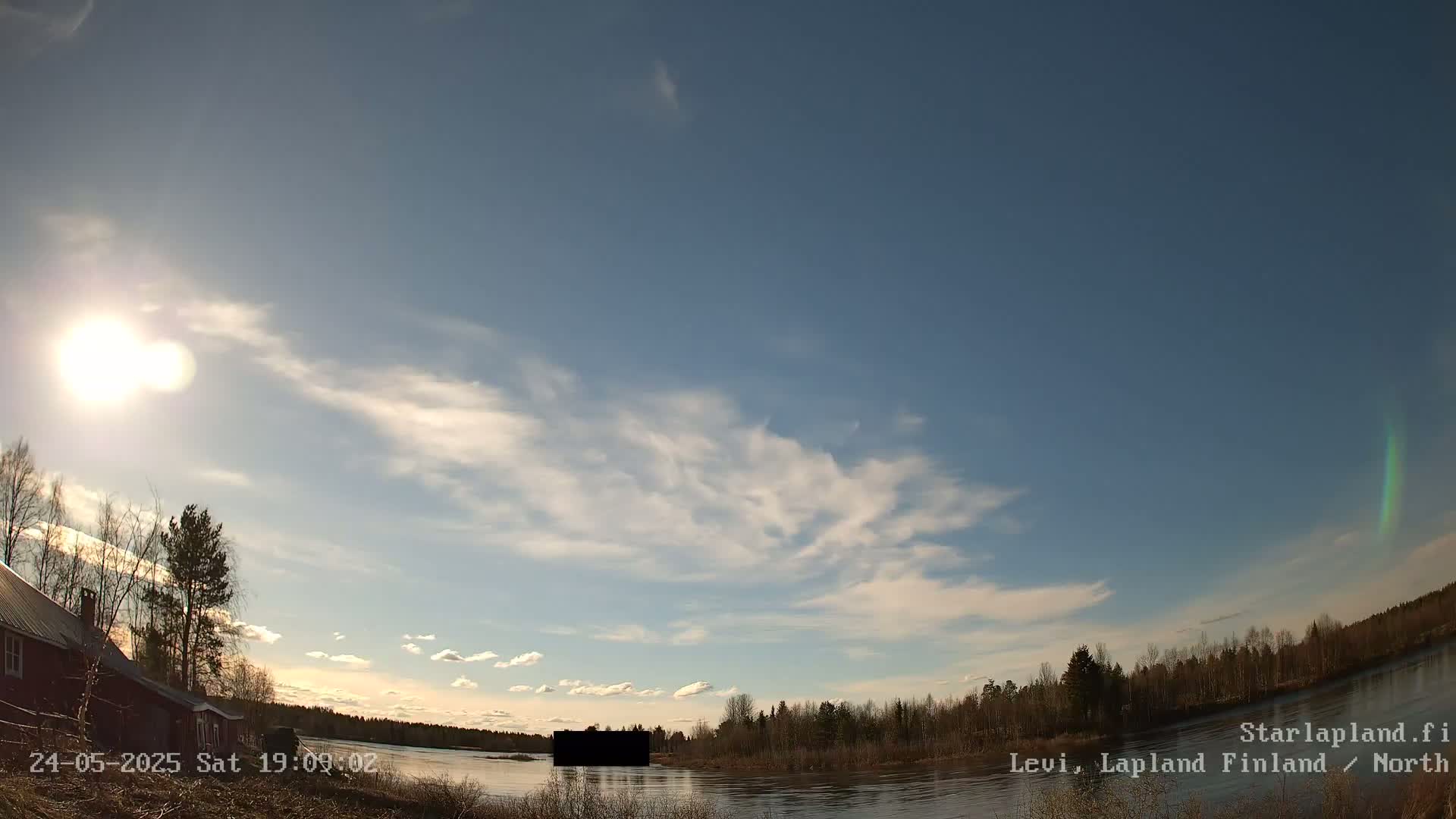 A sunny day with wispy clouds over a calm river and a treeline, with a red building visible near the left edge of the image.