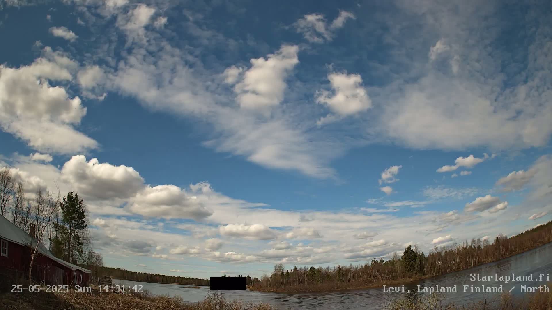 A partly cloudy sky over a calm river, with a red building and trees visible on the left and a treeline on the right.