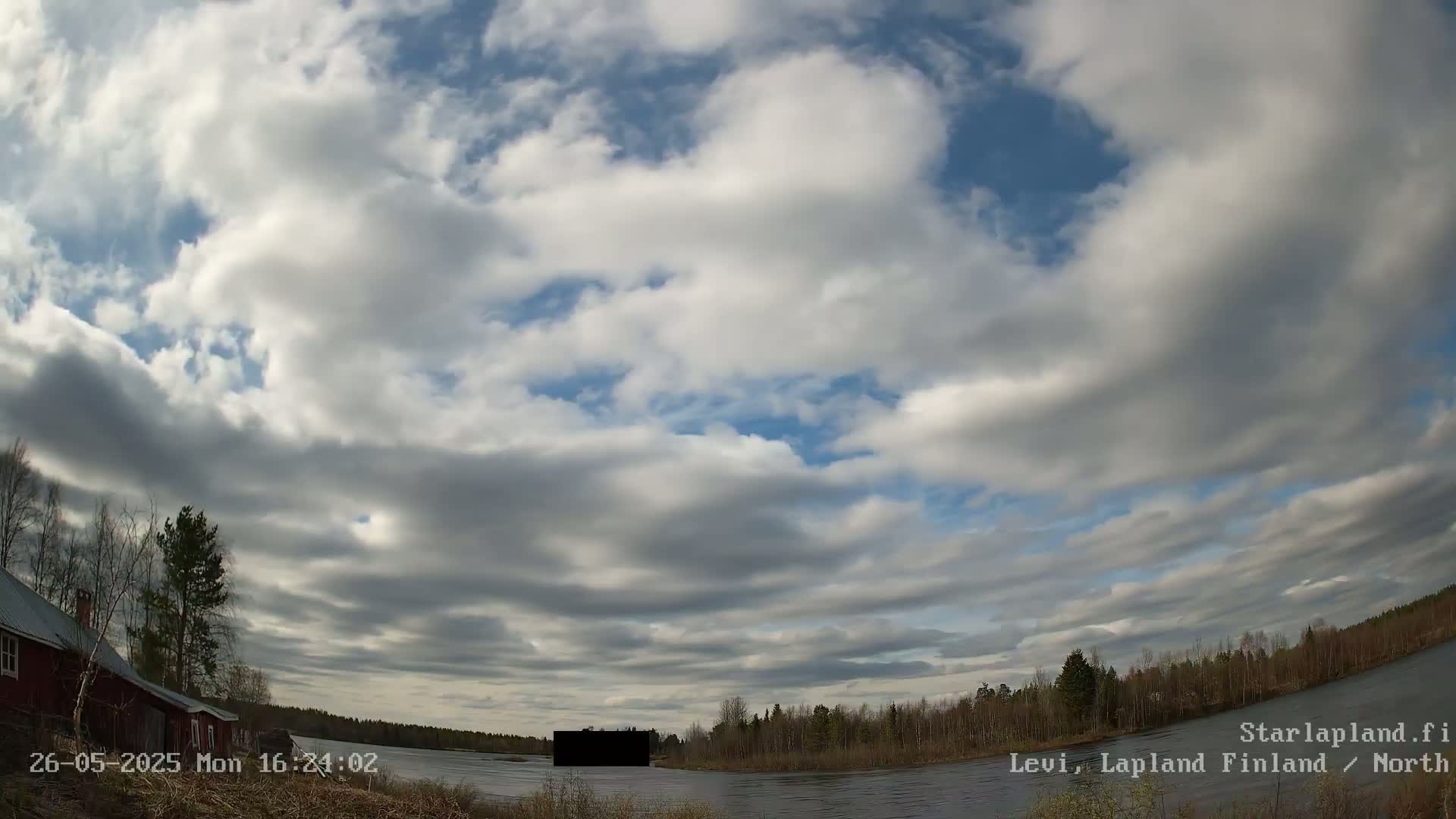 A partly cloudy sky over a calm body of water, with trees and a red building visible on the left.