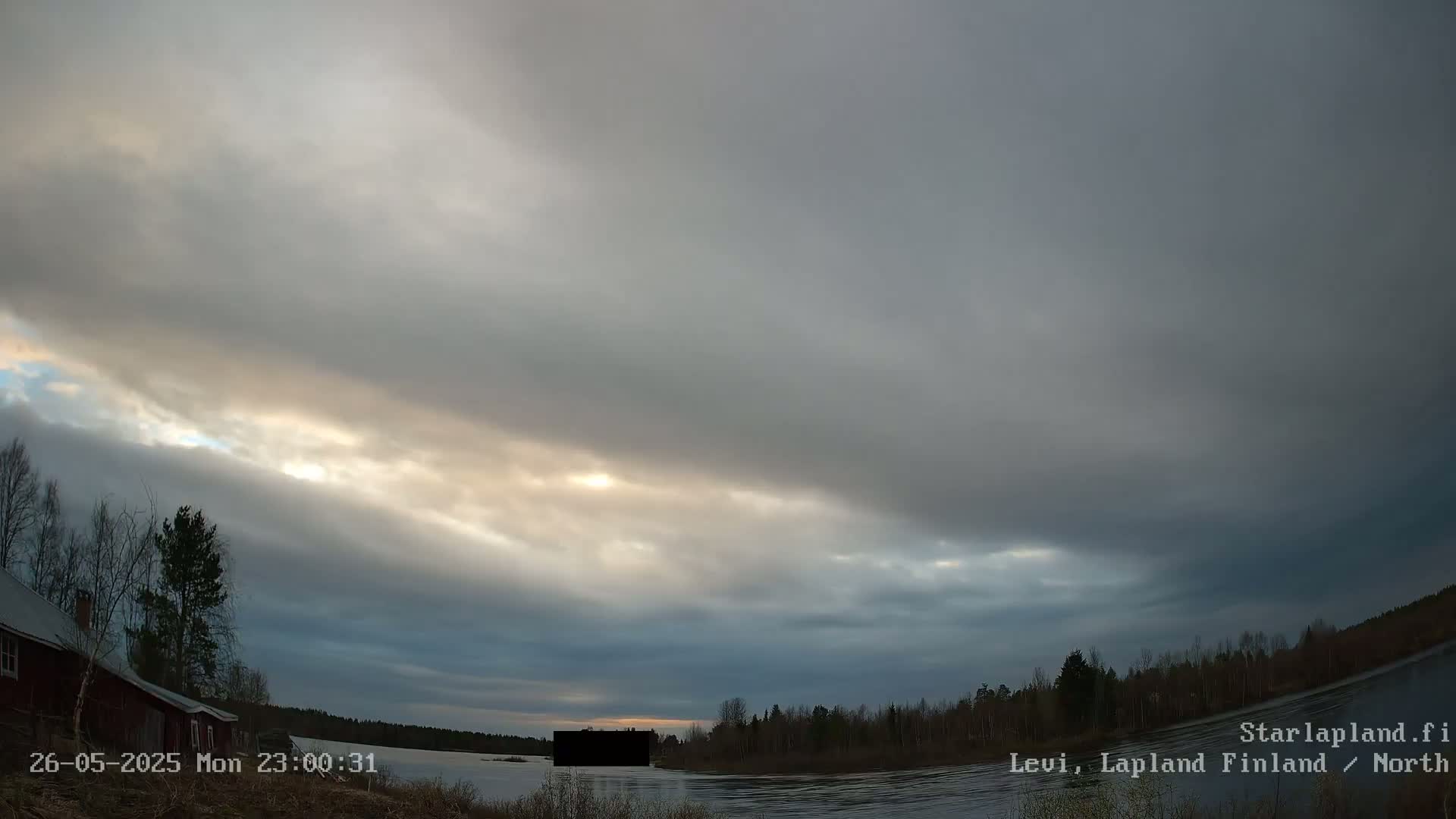 A mostly cloudy sky with some sun breaking through illuminates a calm body of water next to a treeline and a small, dark building.
