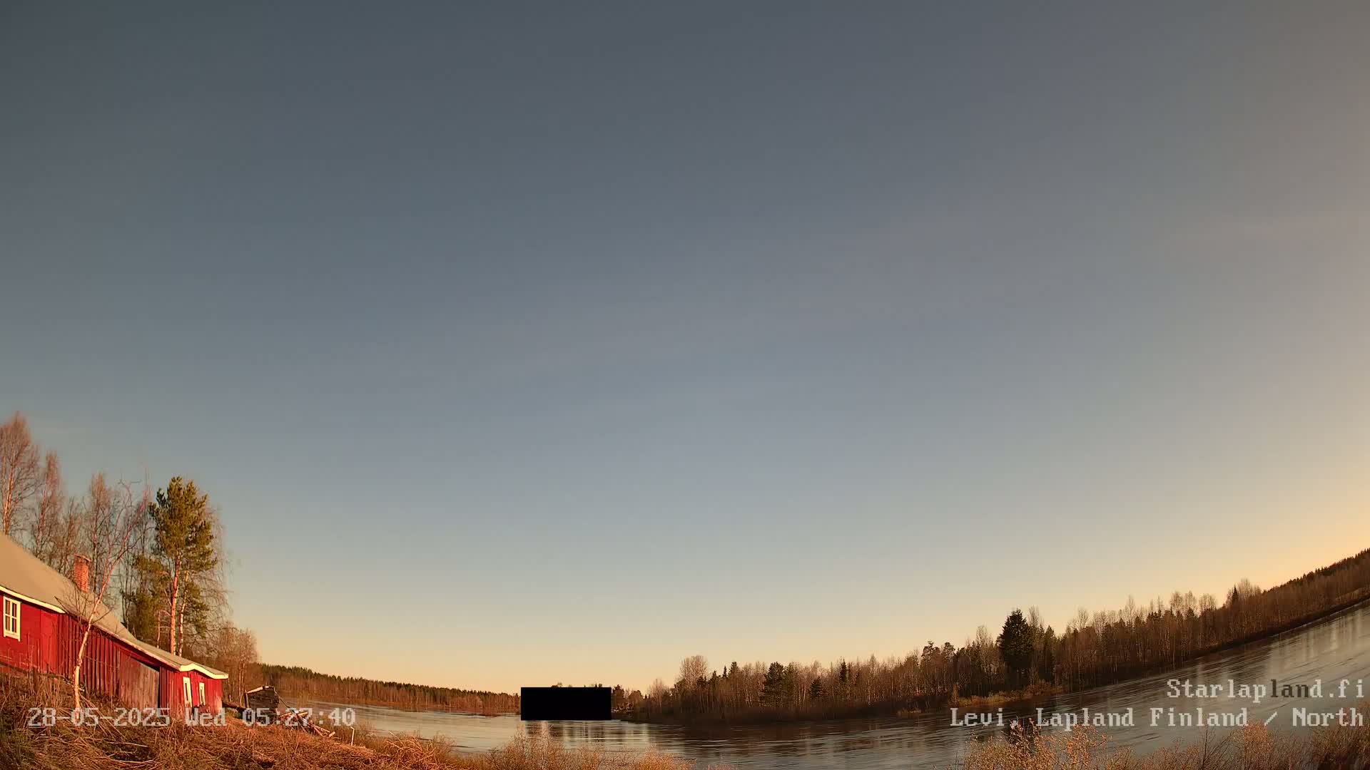 A red house sits near a calm river under a clear, pale sky at sunset, with leafless trees lining the riverbank.