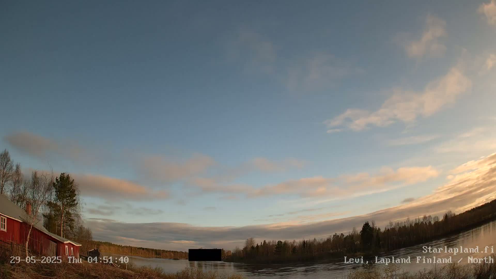 A red house sits near a calm river under a mostly clear sky with pale, wispy clouds at sunrise.