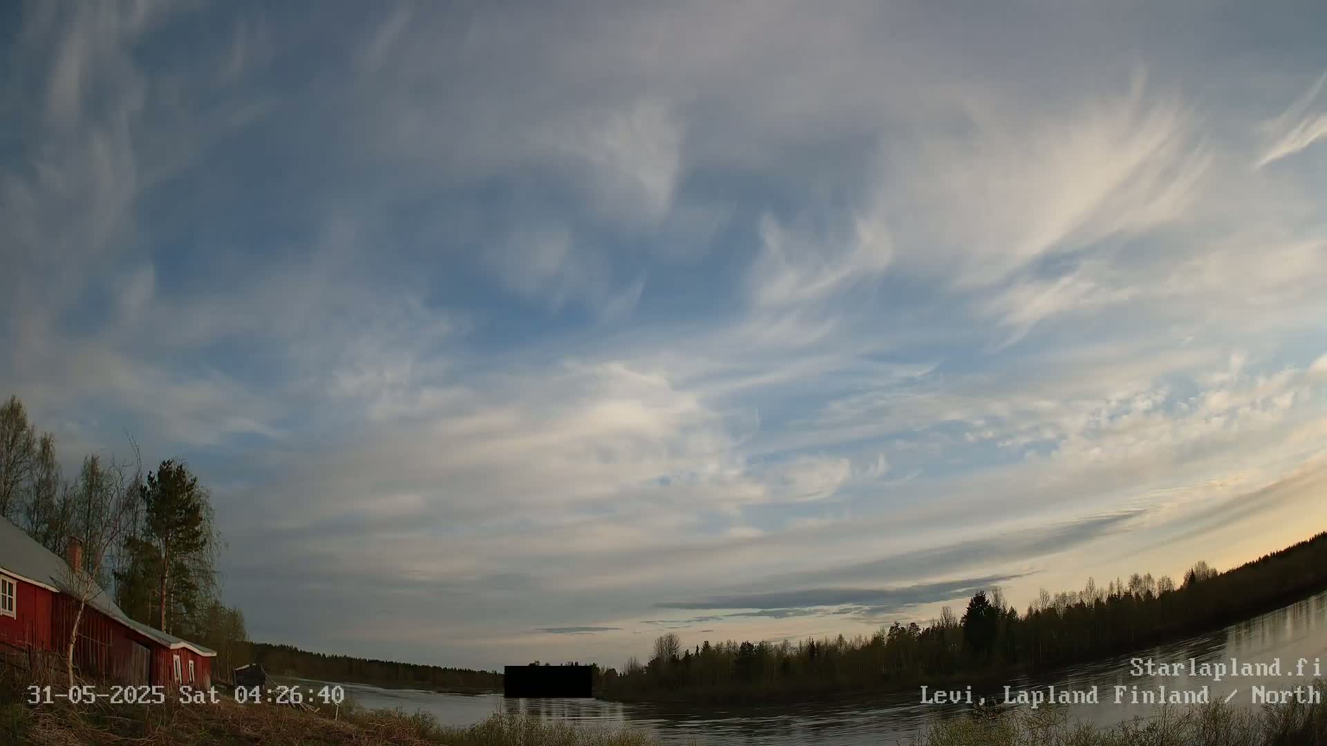 A red house sits near a calm lake under a mostly cloudy sky with wispy cirrus clouds.