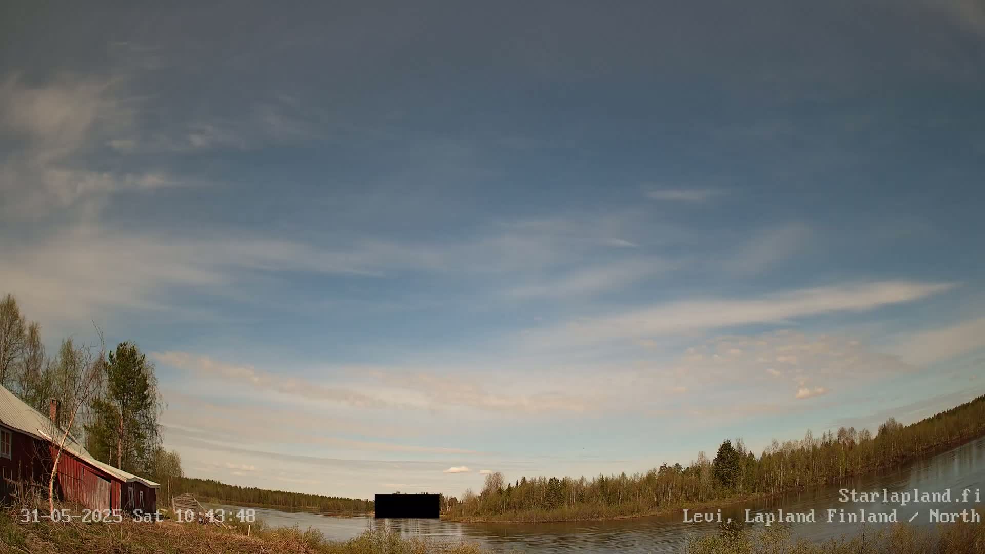 A partly cloudy sky overlooks a calm river bordered by a line of green trees and a small red building.