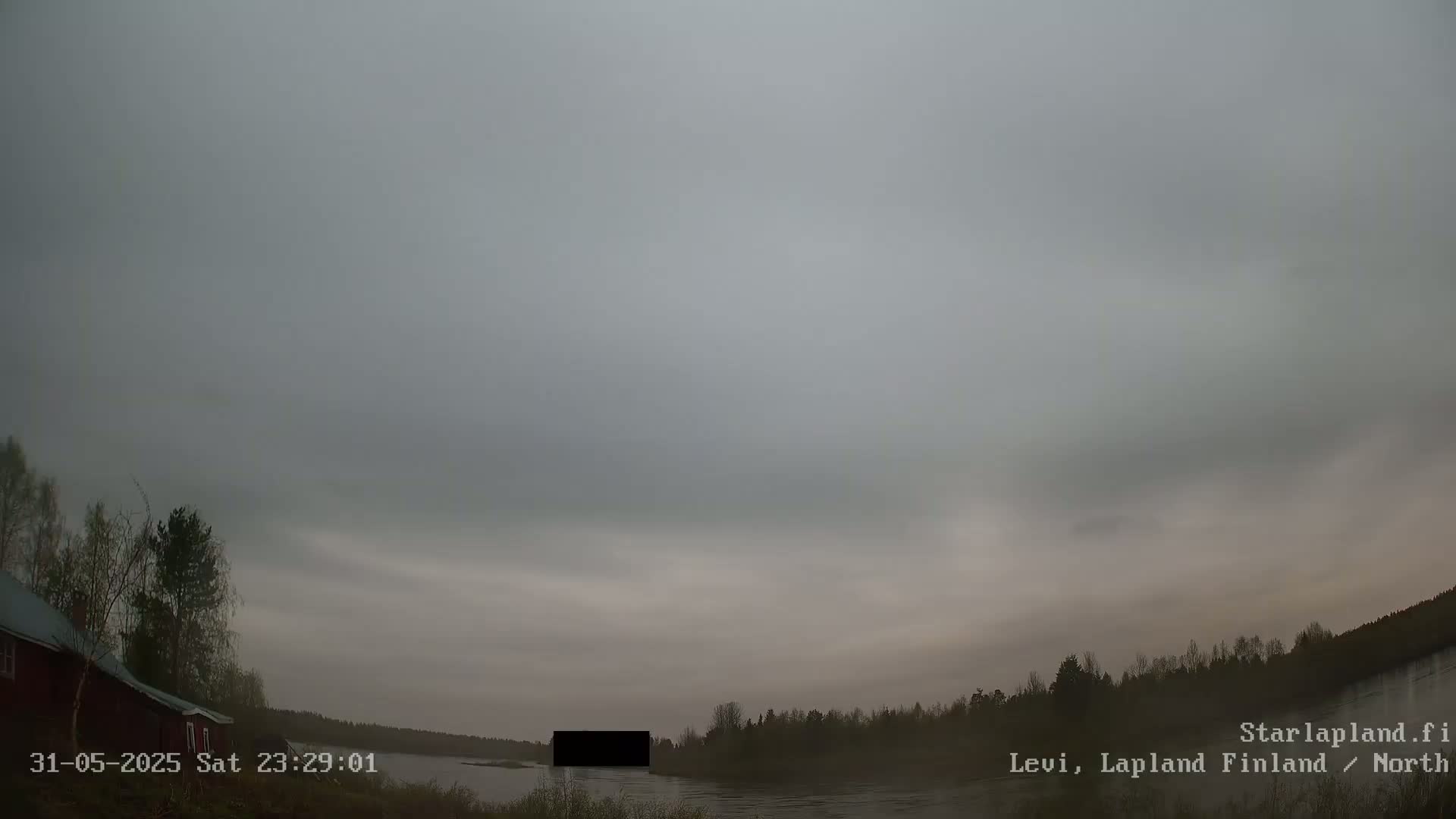 A mostly cloudy, overcast sky hangs over a calm body of water, with dark trees lining the shoreline and a red building visible on the left.