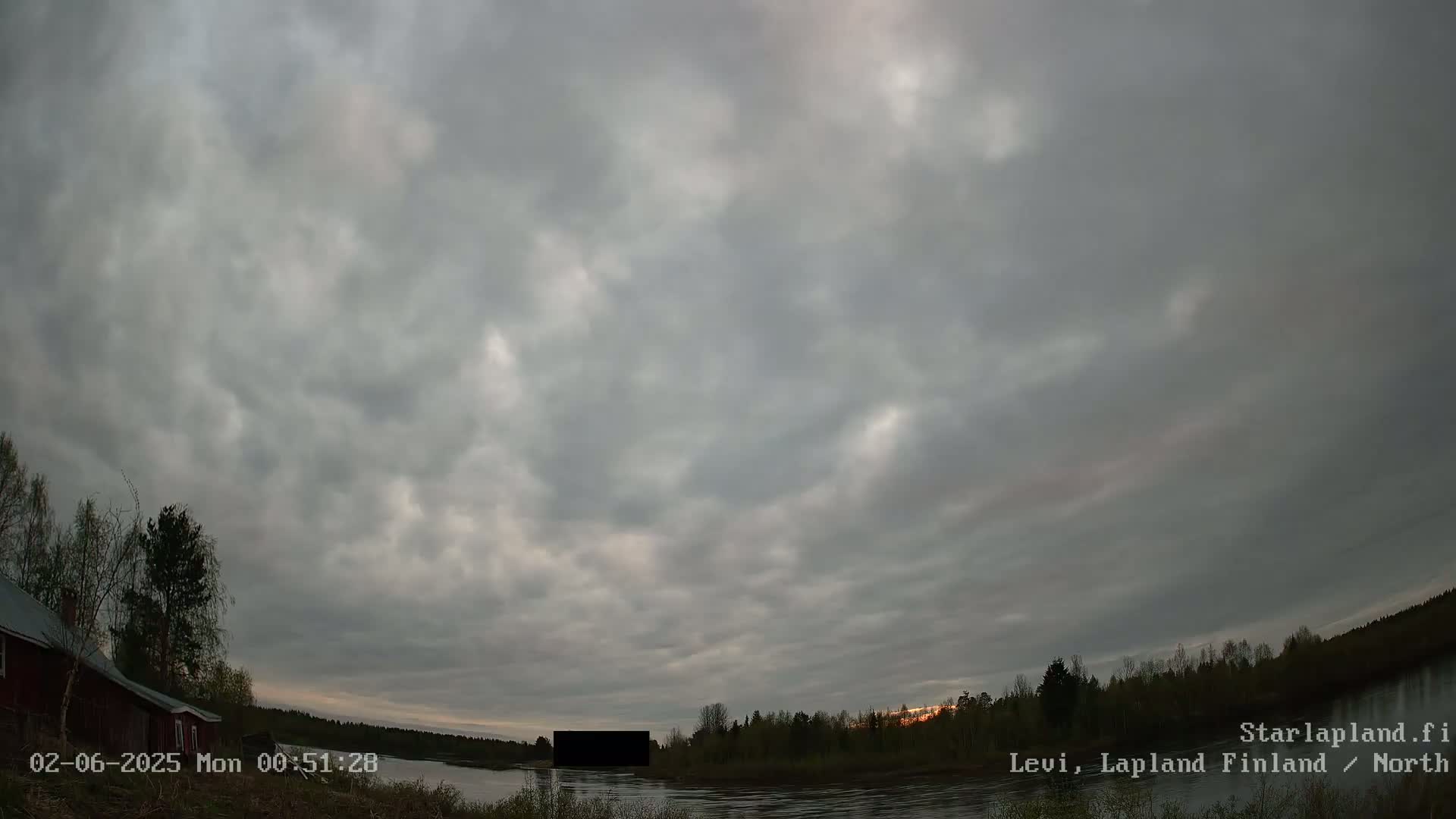 A mostly cloudy twilight sky over a calm body of water, with a red building and trees visible on the left bank.