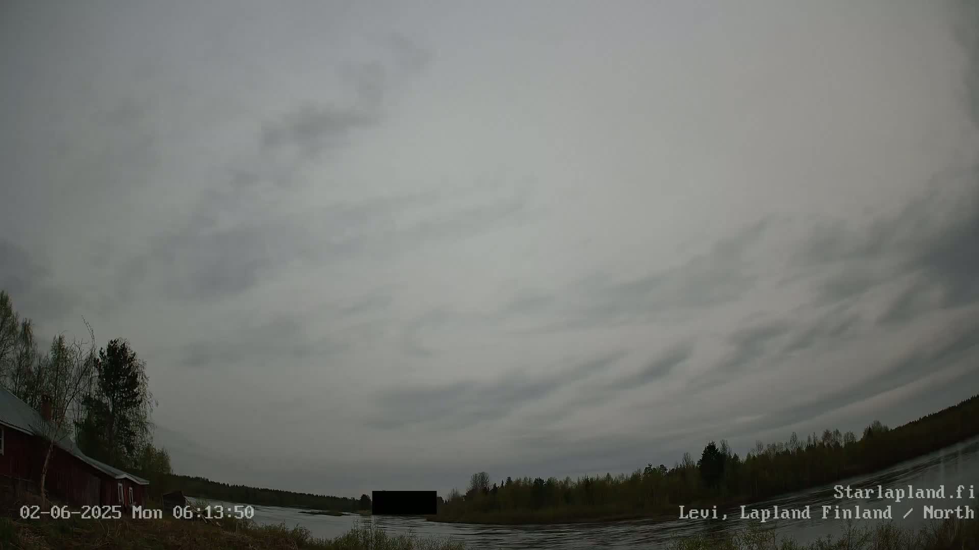 A mostly cloudy sky hangs over a calm river, bordered by trees and a small red building.
