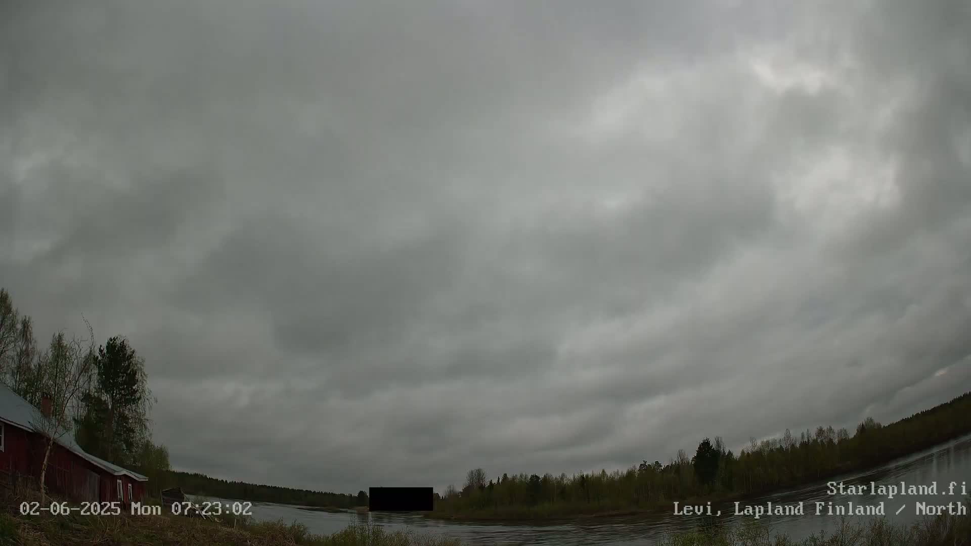 A wide shot of an overcast sky over a calm river, with trees and a red building visible along the shoreline.