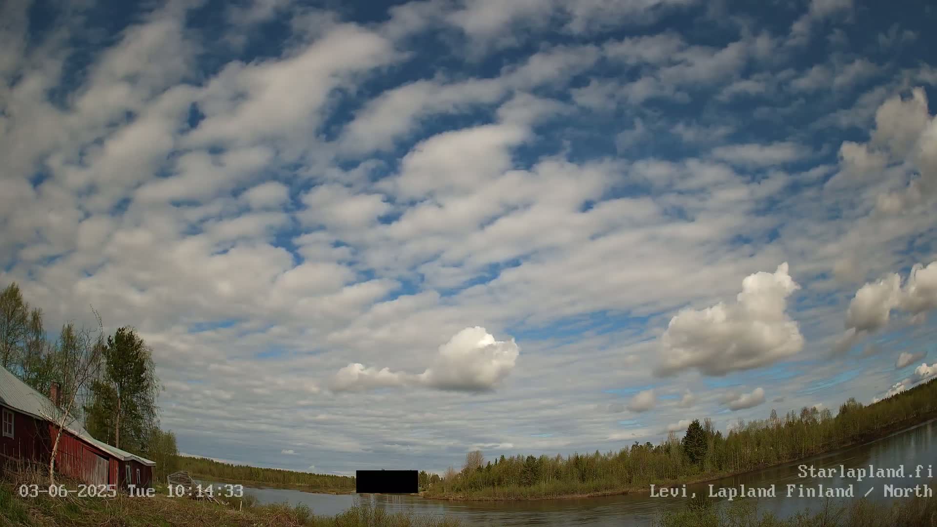 A mostly sunny day with scattered cumulus clouds over a calm river, showing a red building on the left and a dark structure on the water.