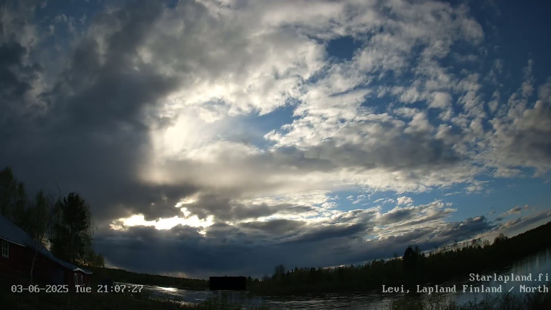 A partly cloudy sky with breaks of sunlight over a calm body of water and a treeline, near a red building.
