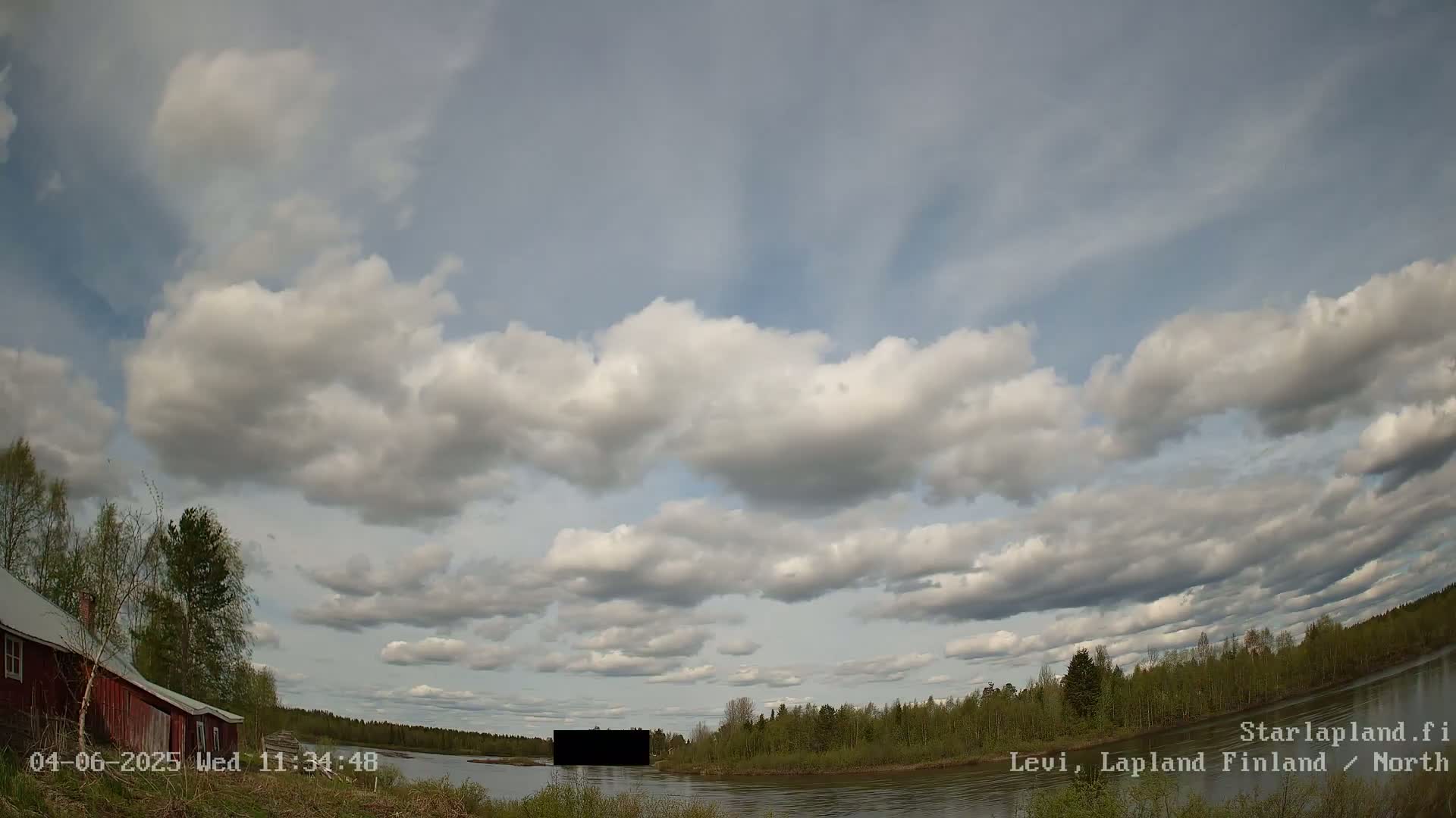 A partly cloudy sky over a calm river, with a red building and green trees visible on the shore.