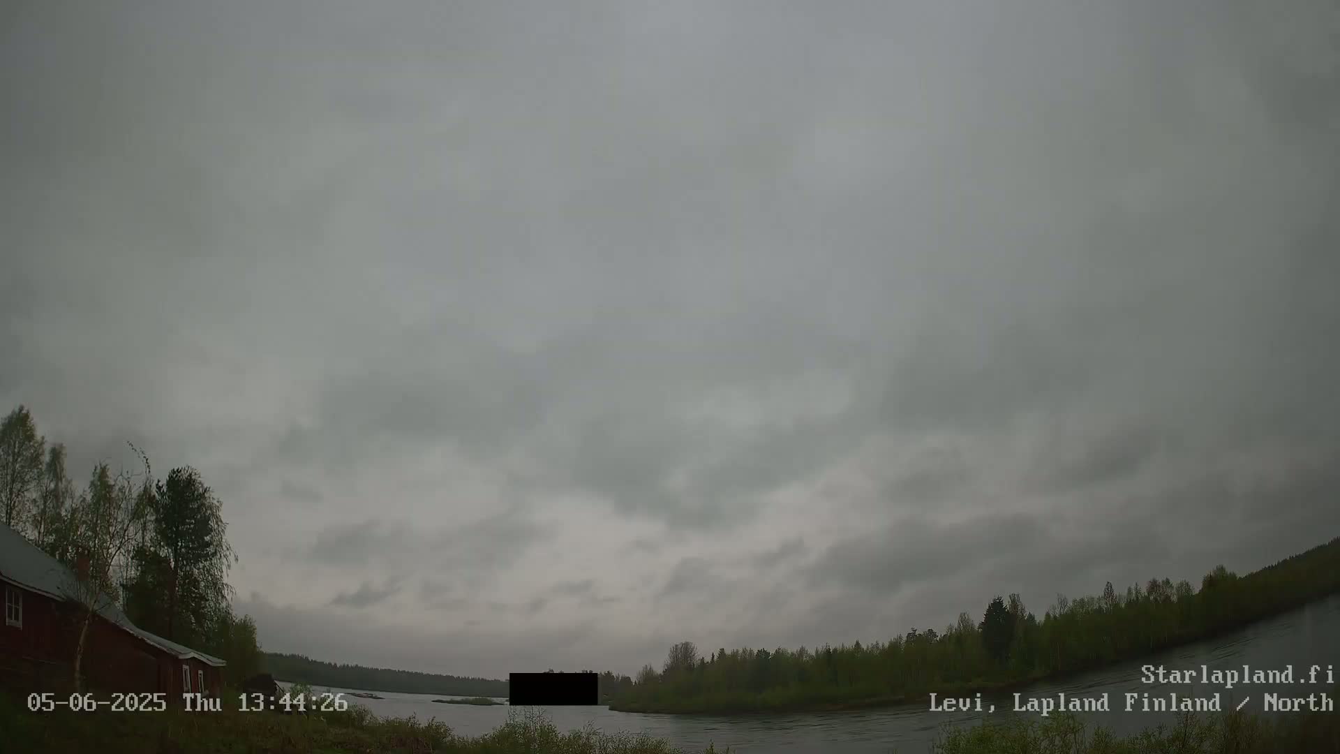 A gray, overcast sky hangs over a calm river bordered by dark green trees and a red building on the left.