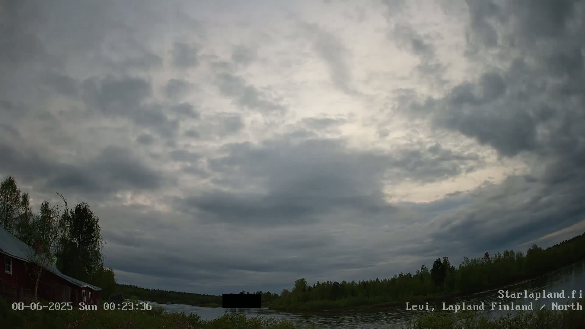 A cloudy, overcast sky hangs over a calm river that winds through a dark green, forested shoreline, near a small red building.