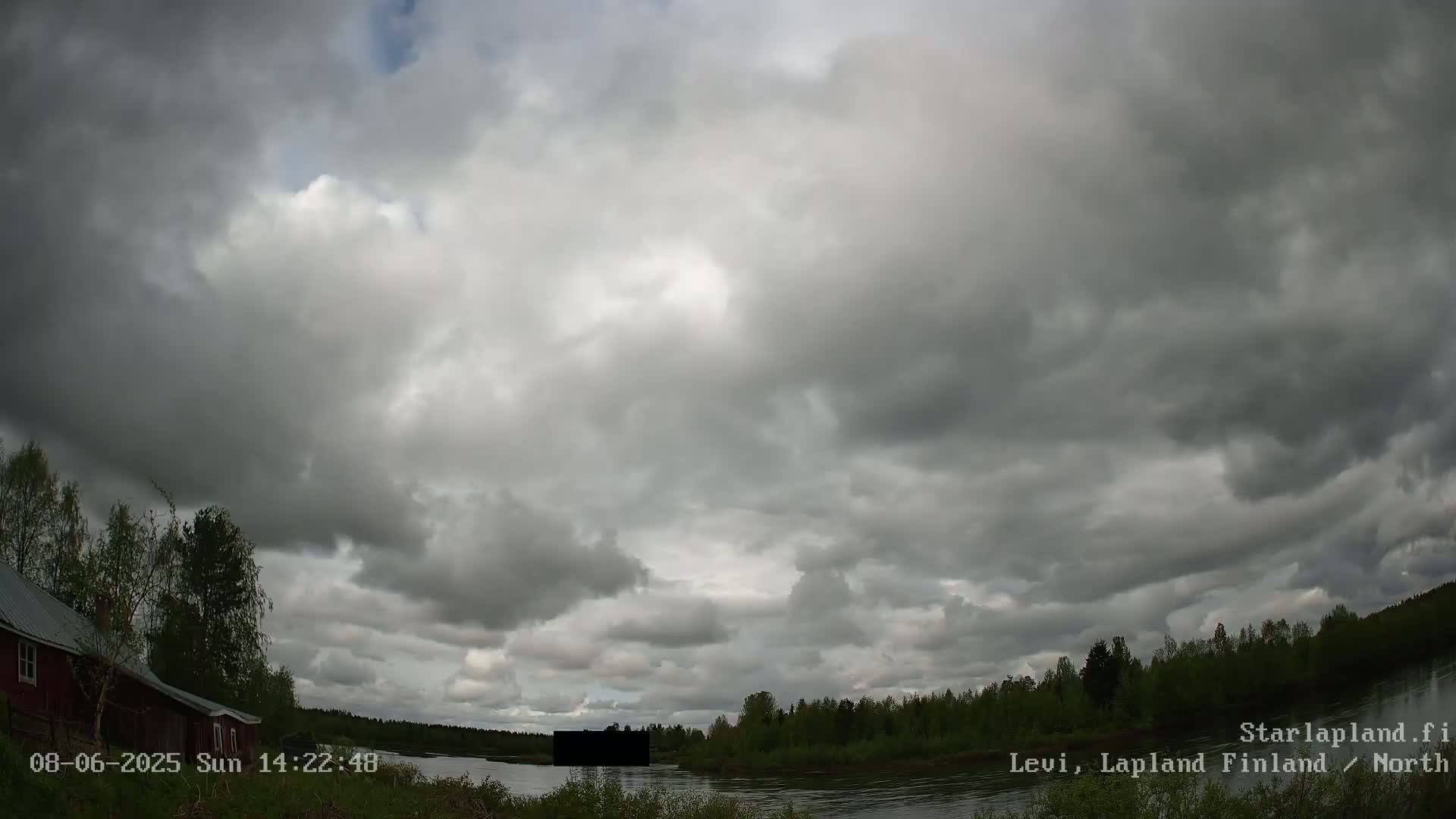 A cloudy sky hangs over a calm river, bordered by green trees and a red building on the left.