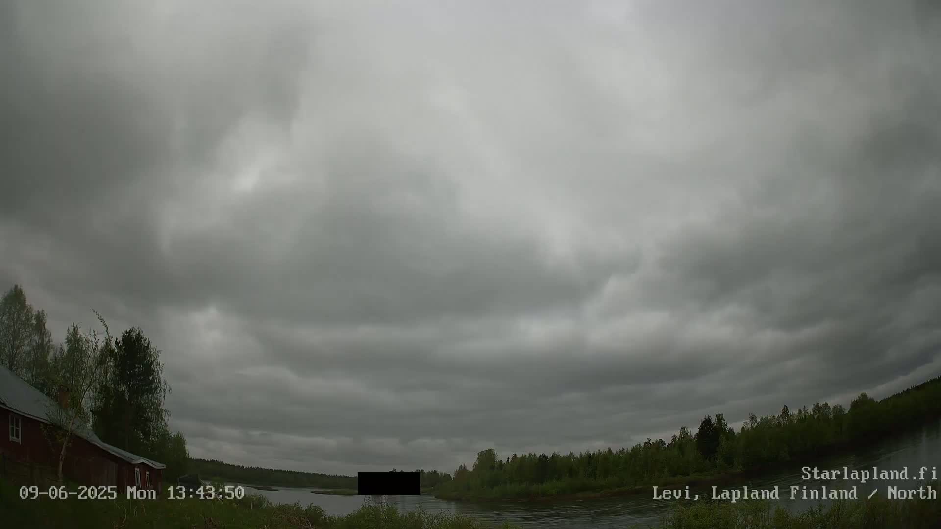 A wide shot shows an overcast sky over a river, with trees and a red building visible on the left bank.