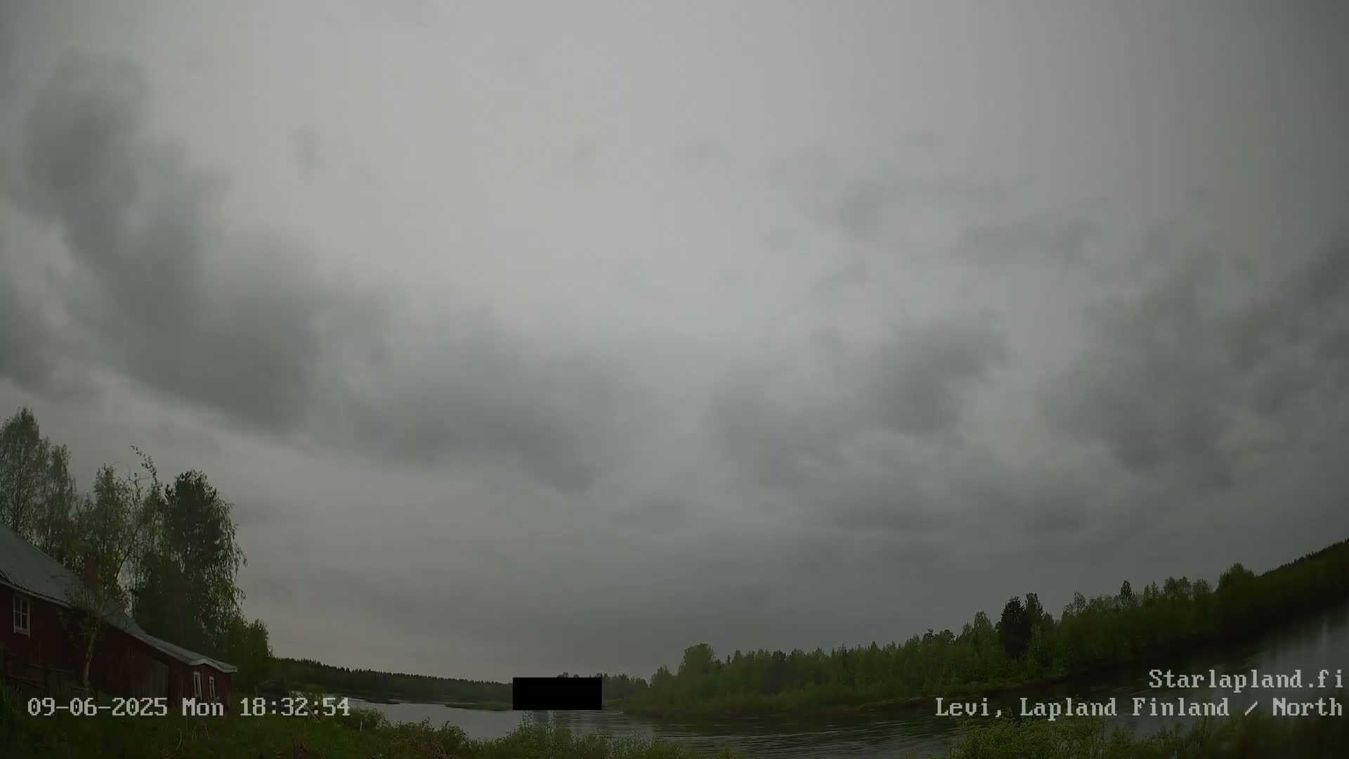A gray, overcast sky hangs over a calm river bordered by a dark green forest and a red building.