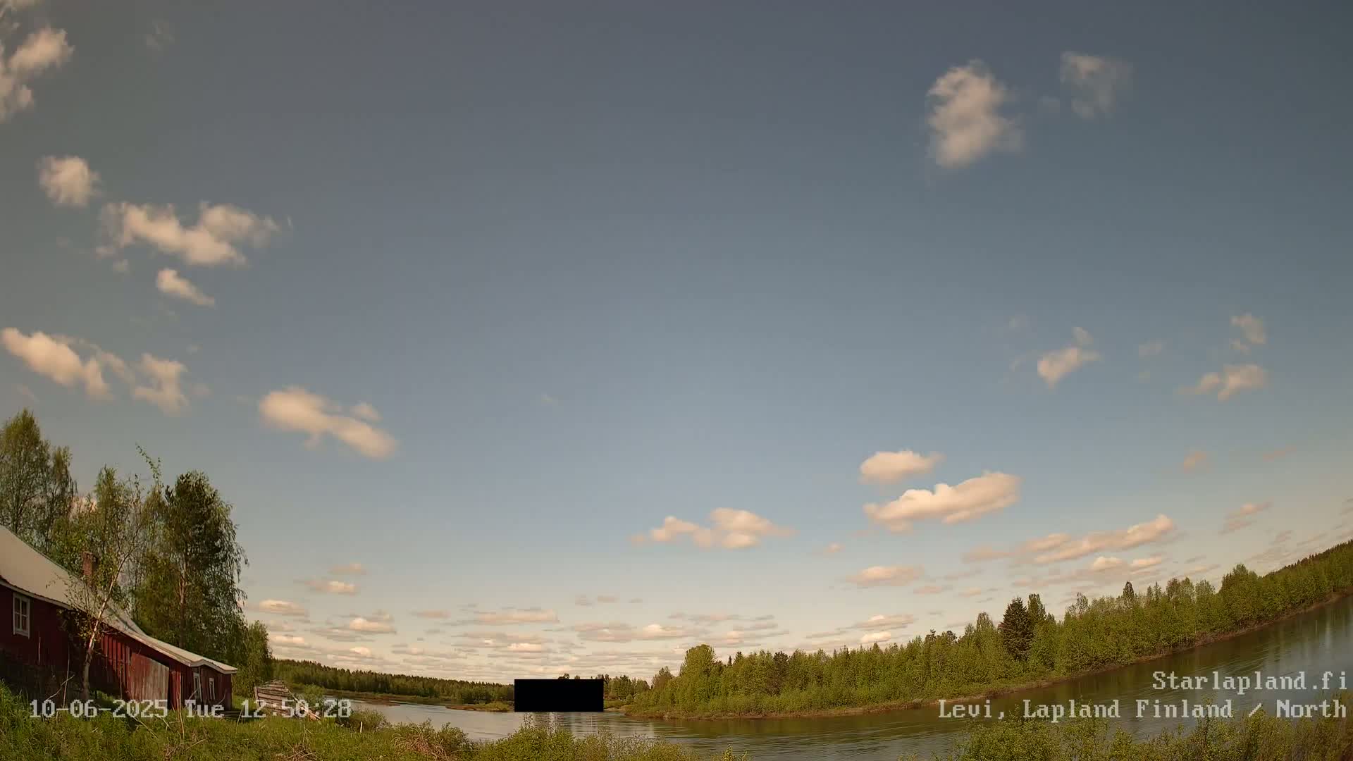 A red building sits near a calm river bordered by lush green trees under a partly cloudy sky.