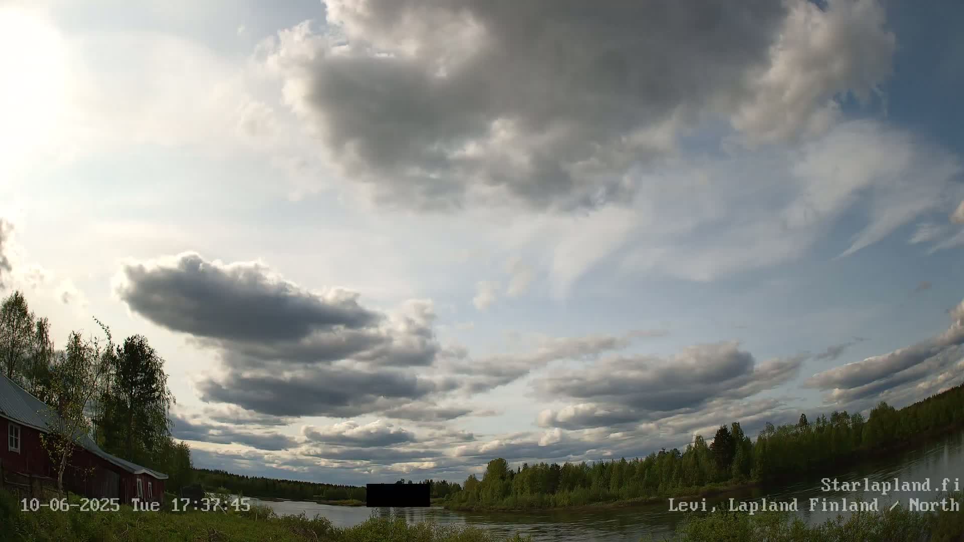 A partly cloudy sky hangs over a calm river bordered by green trees and a red building.