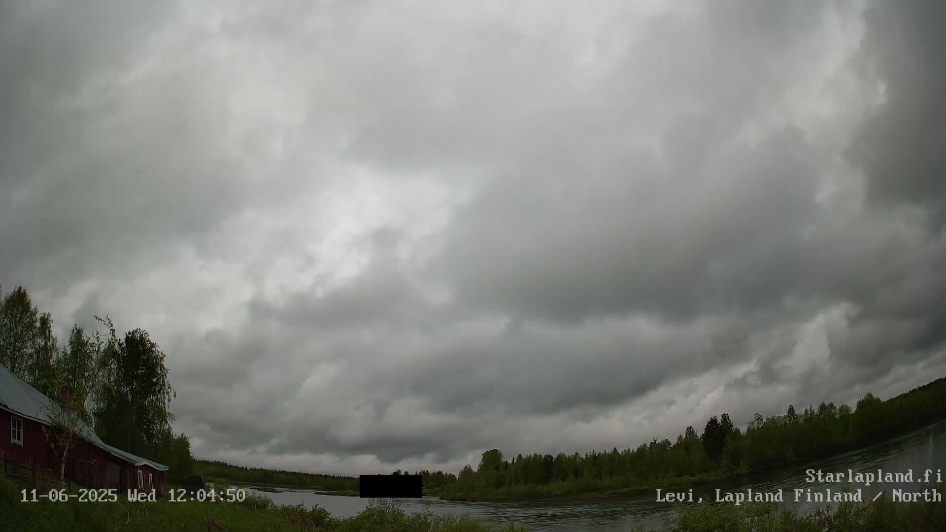 A cloudy, overcast sky hangs over a calm river and forested shoreline, with a red building visible near the riverbank.