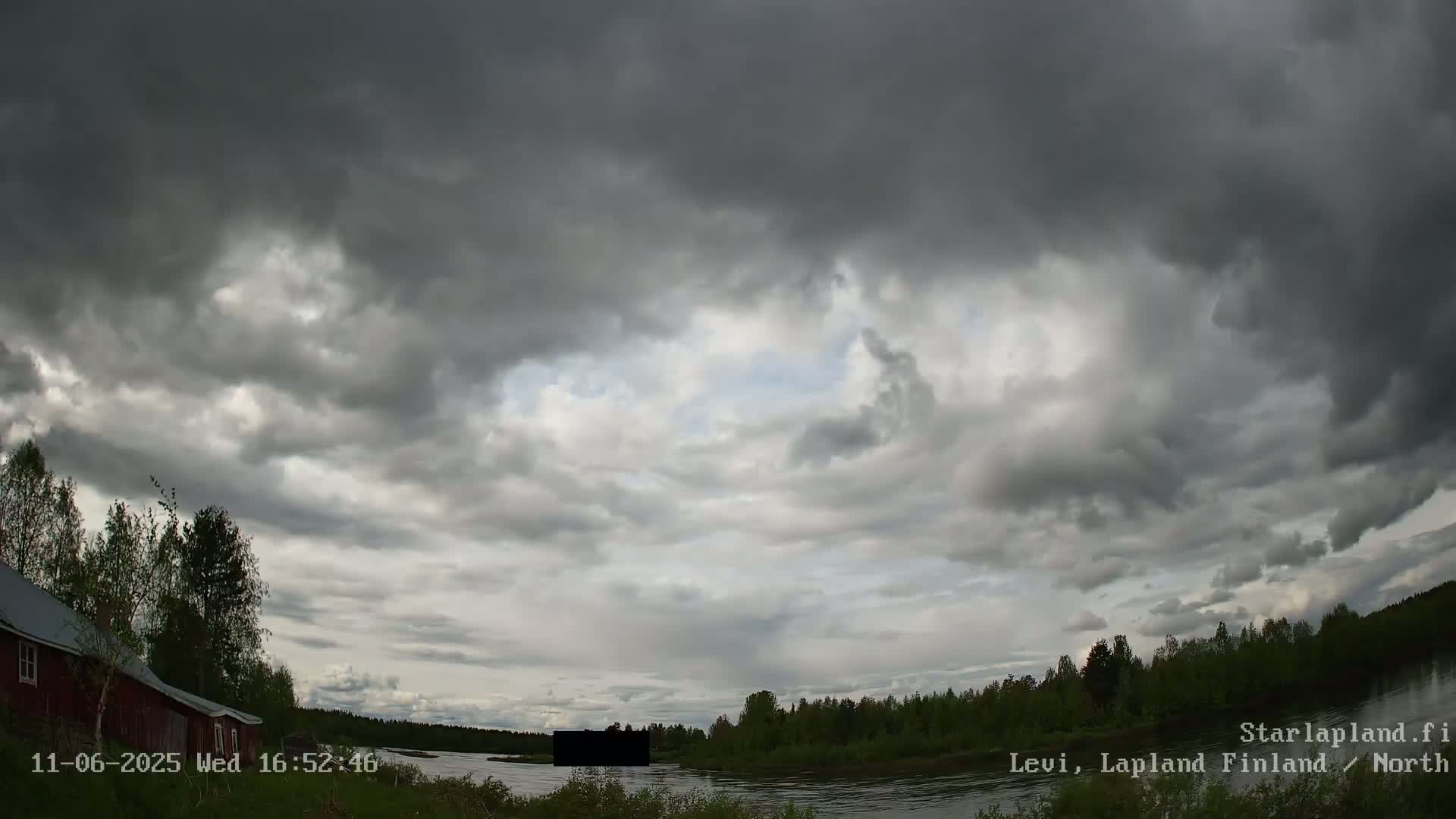 Overcast skies loom over a calm river, bordered by a treeline and a small red building.