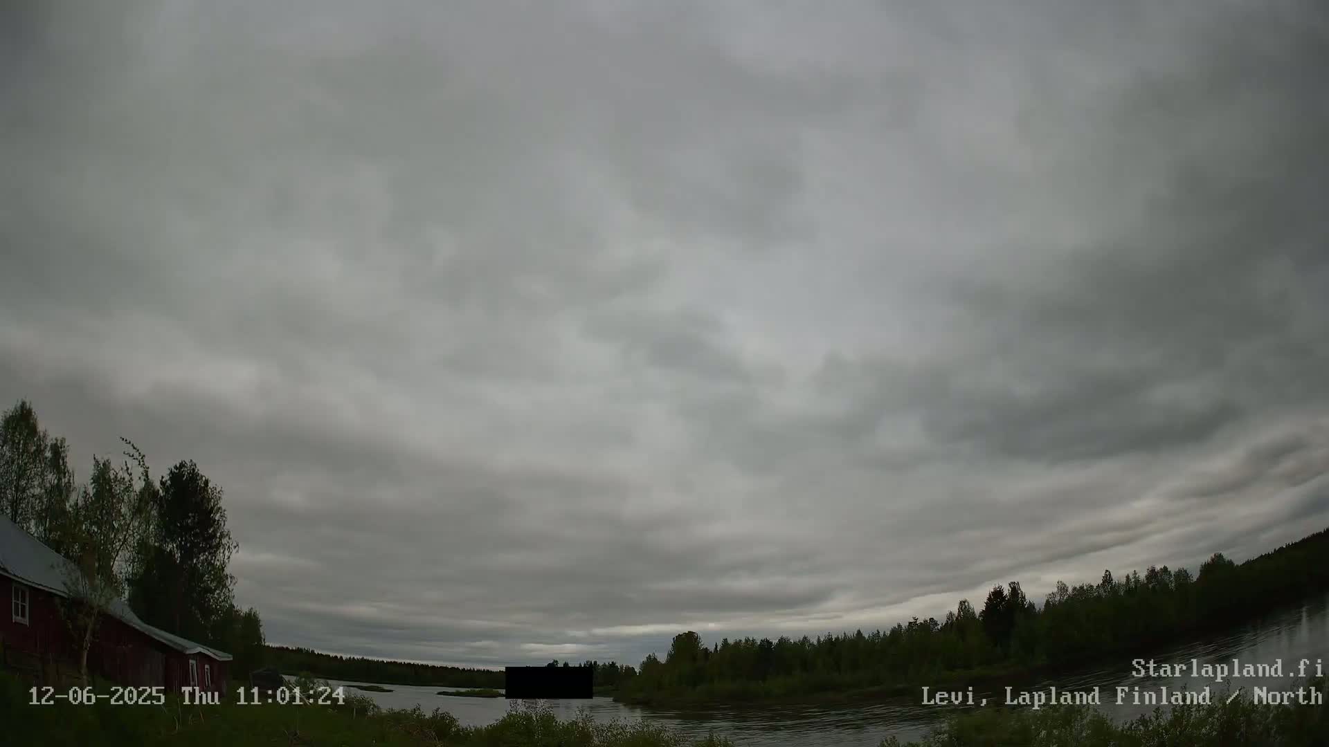 A mostly cloudy sky hangs over a calm river bordered by dark green trees and a red building.