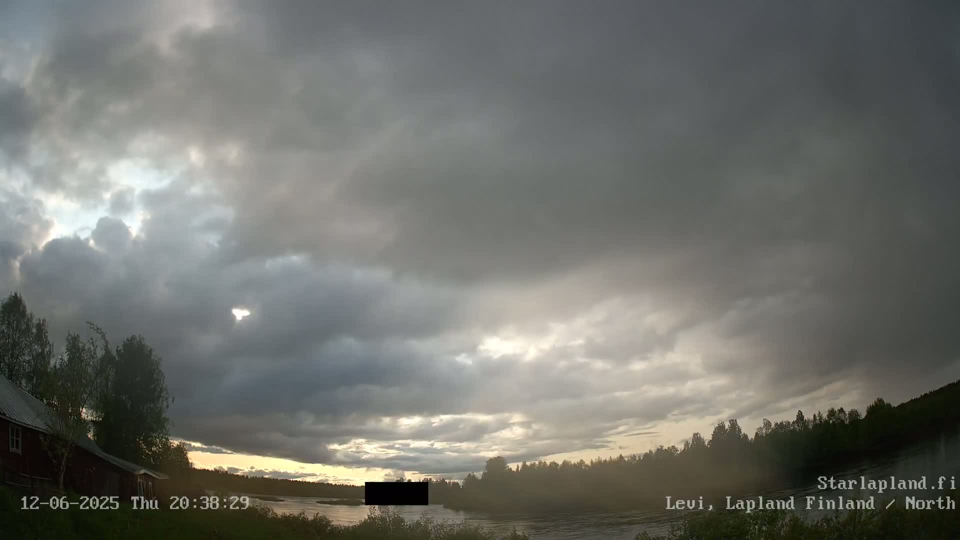 A mostly cloudy sky with some sunlight breaking through over a calm lake and forested shoreline, near a small red building.