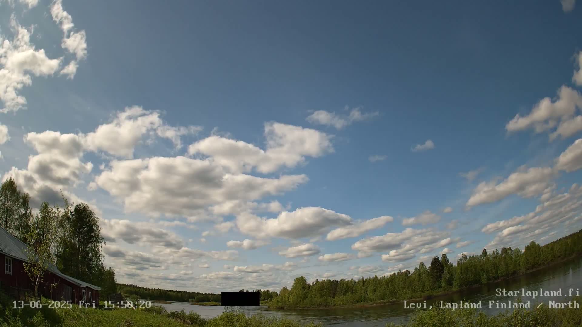 A partly cloudy sky over a calm river, with green trees lining the banks and a red building visible on the left.