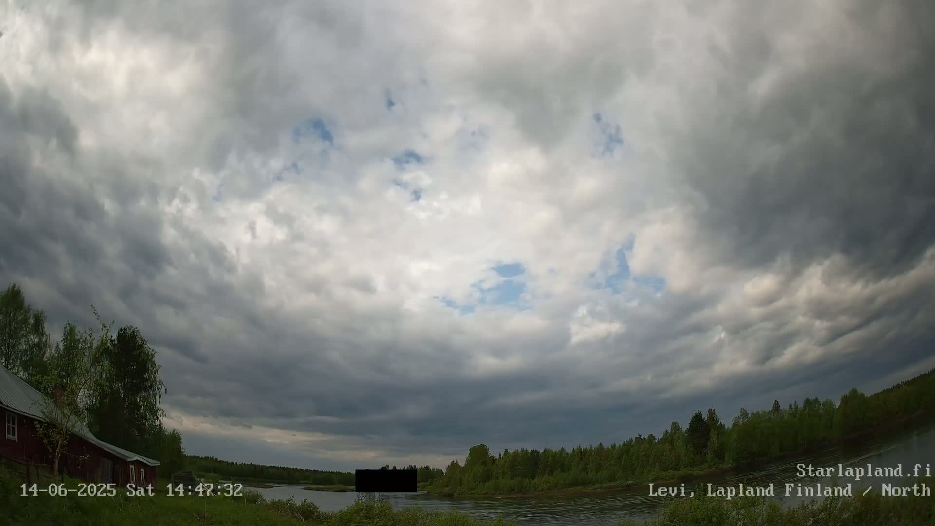 A mostly cloudy sky hangs over a calm river bordered by green trees and a red house.