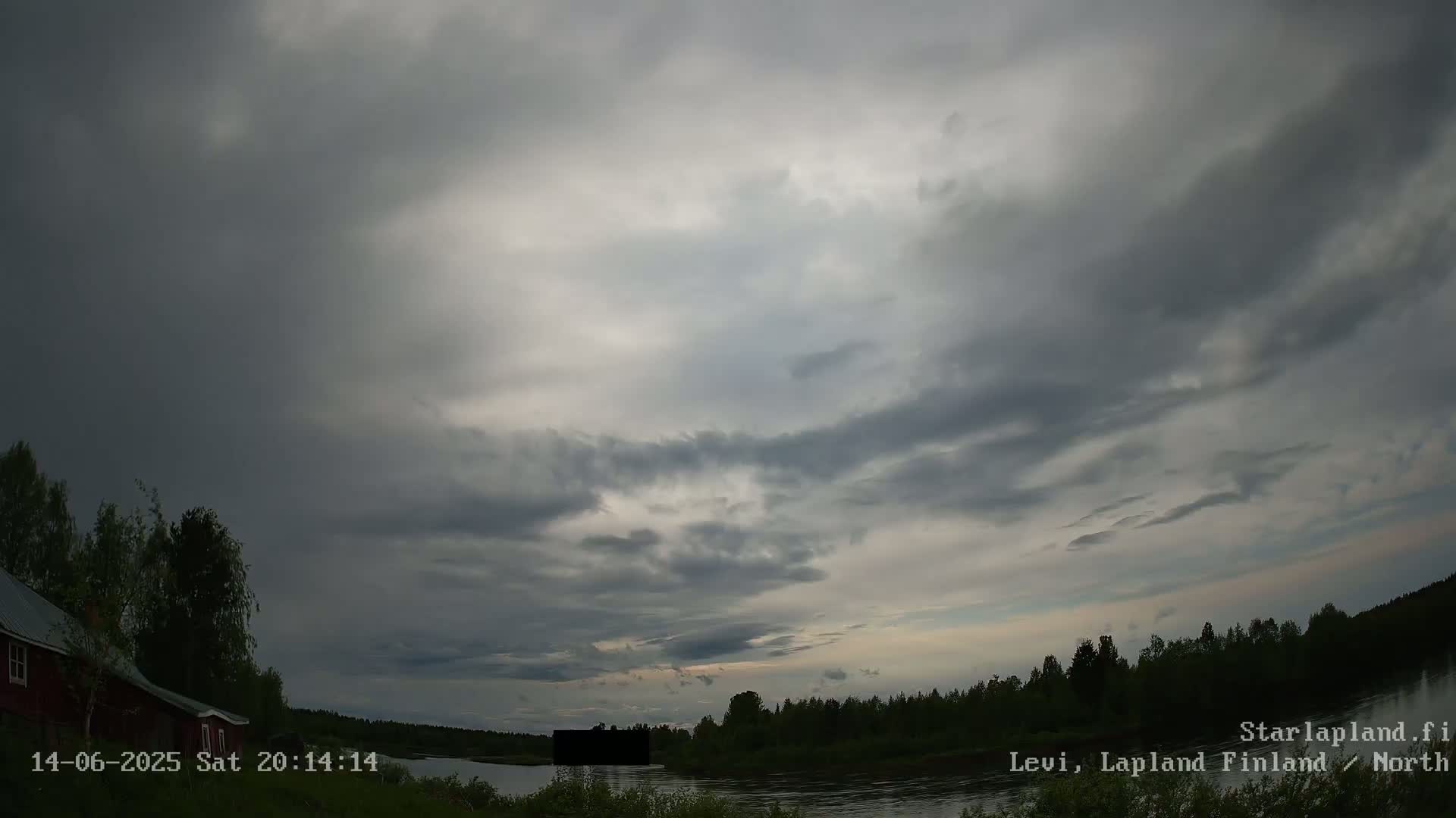 A mostly cloudy, overcast sky hangs over a calm river bordered by dark trees and a small red house.