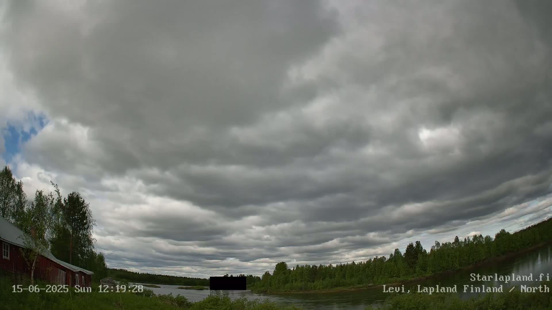 A wide shot shows an overcast sky over a calm river, bordered by a line of green trees and a small red house.