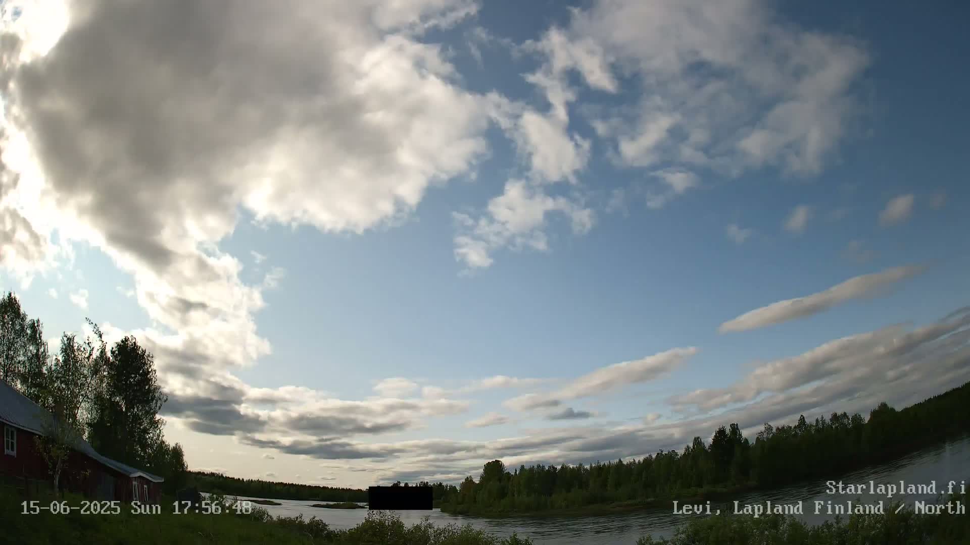 A partly cloudy sky over a calm body of water, with a red building and green trees visible on the shoreline.