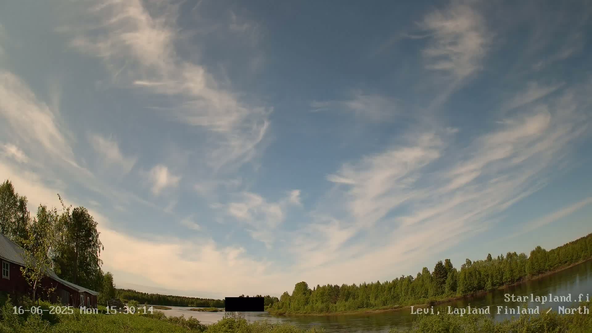 A mostly clear, blue sky with wispy cirrus clouds above a calm river bordered by green trees and a red building.
