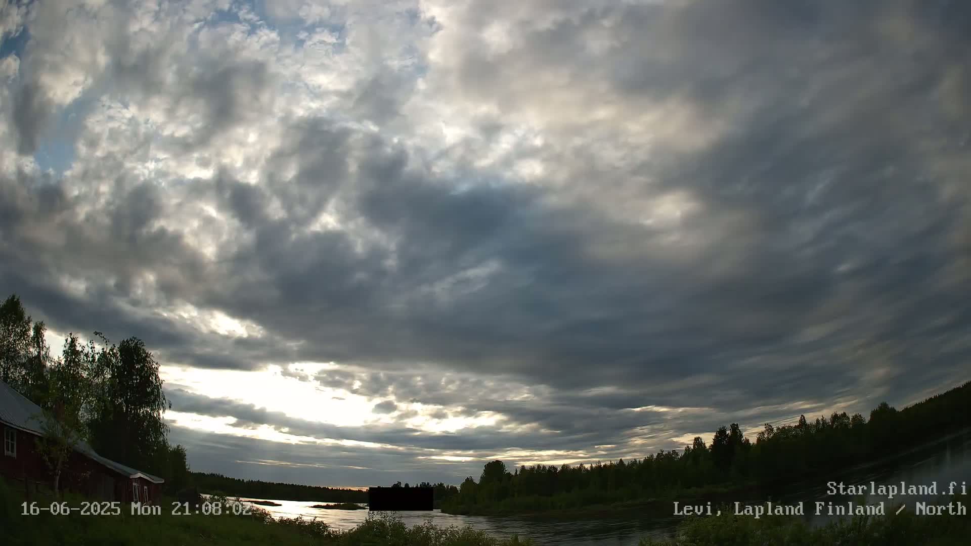 A mostly cloudy sunset over a calm lake and a treeline, with a small building visible on the left.