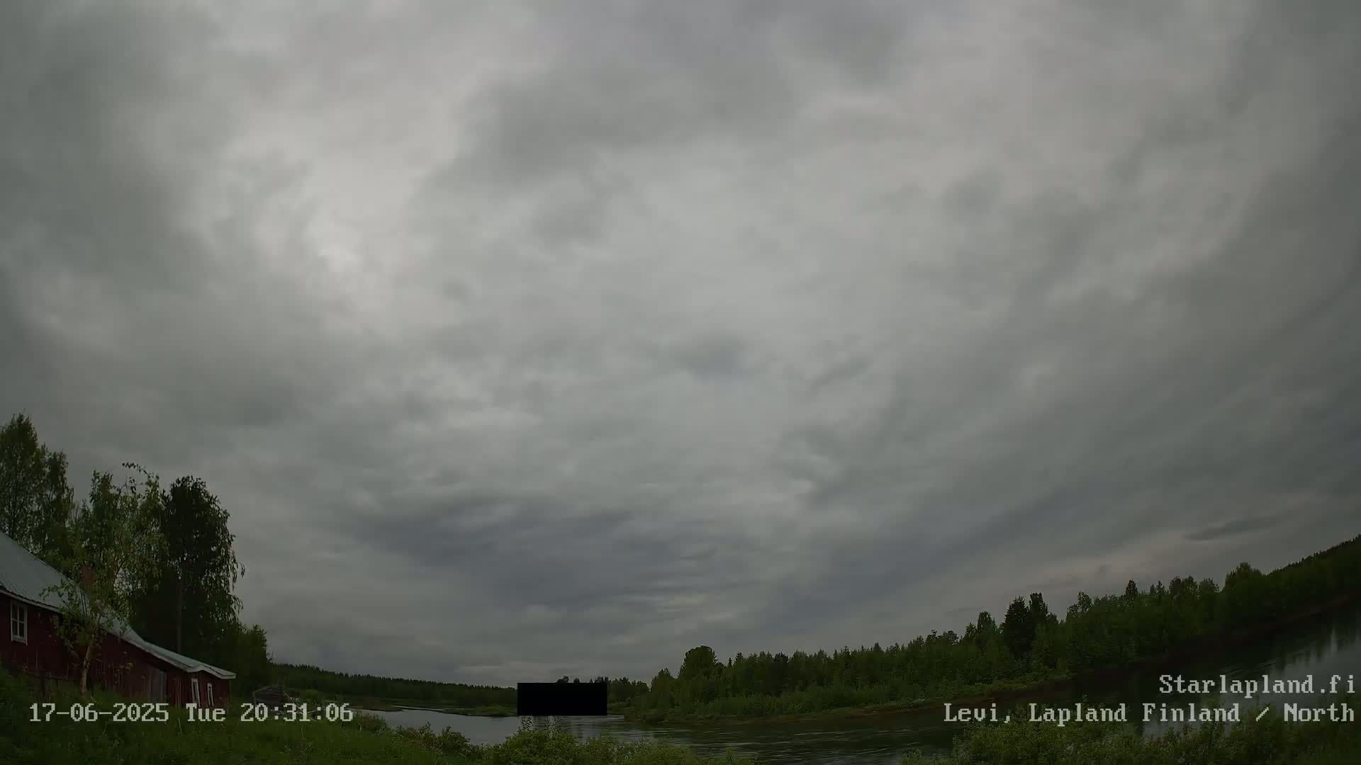 A dark, overcast sky hangs over a calm river bordered by a line of trees and a red building to the left.