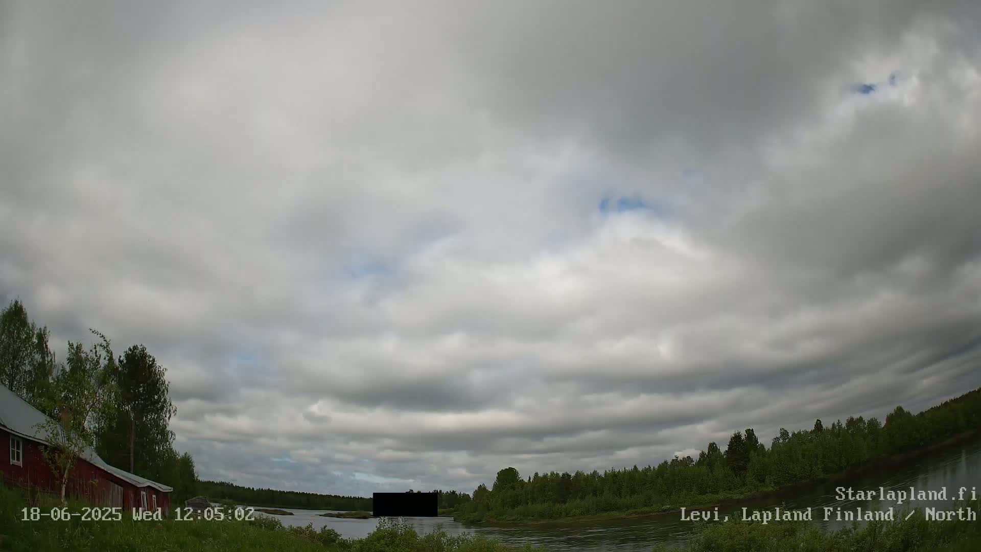 A mostly cloudy sky hangs over a calm river bordered by green trees and a red building to the left.