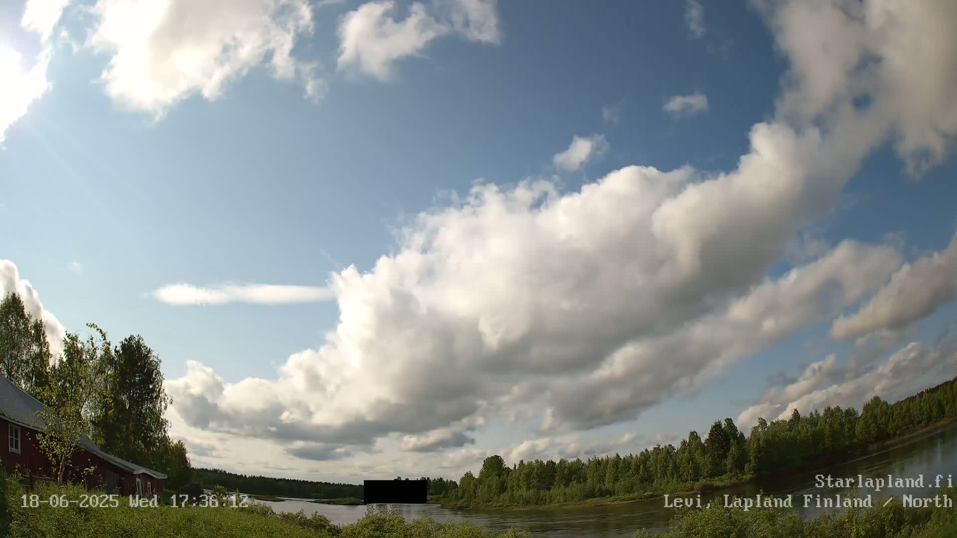 A partly cloudy sunny day shows a river bordered by green trees and a red building on the left.