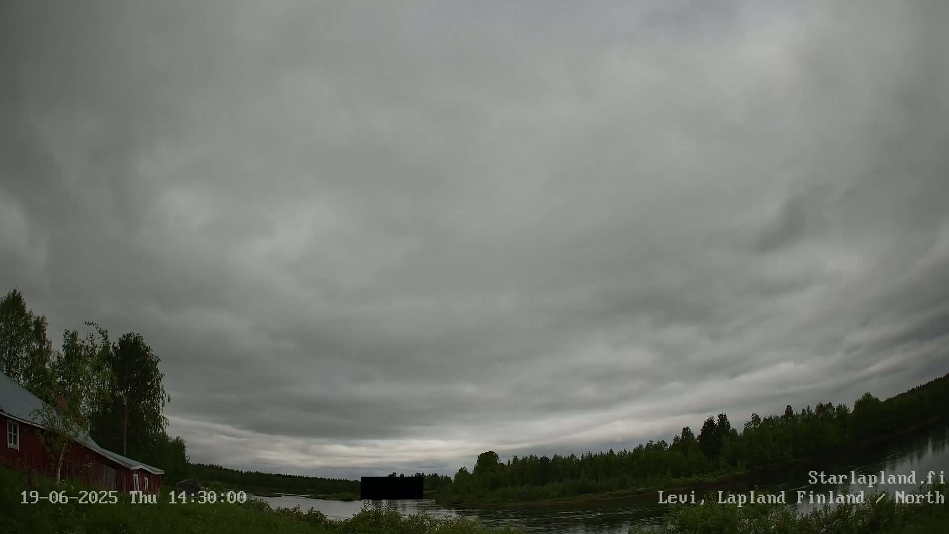 A red building sits near a calm river under an overcast sky, with green trees lining the riverbank.