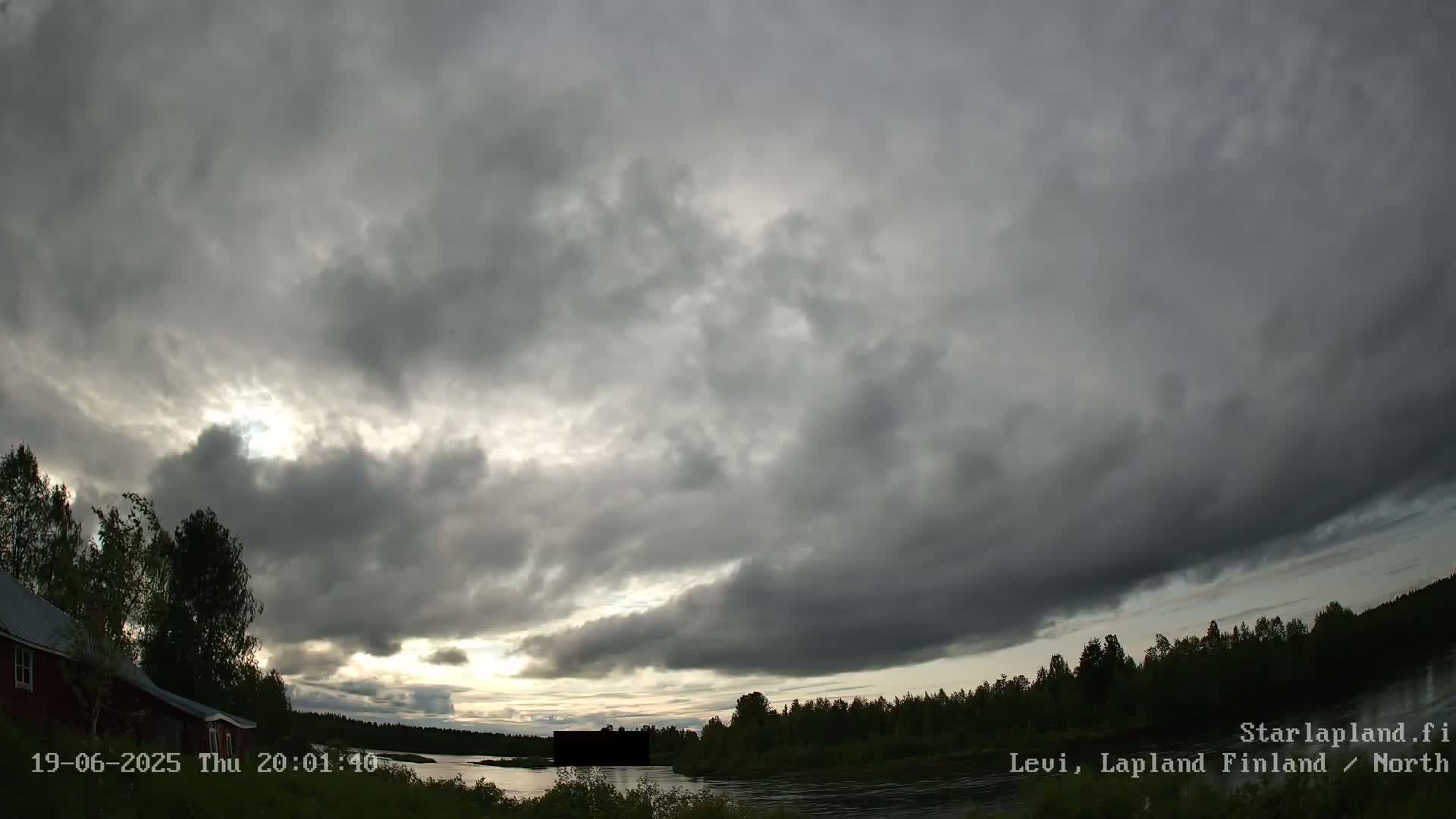 A mostly cloudy sky with some sunlight breaking through over a calm river, bordered by dark green trees and a red building.