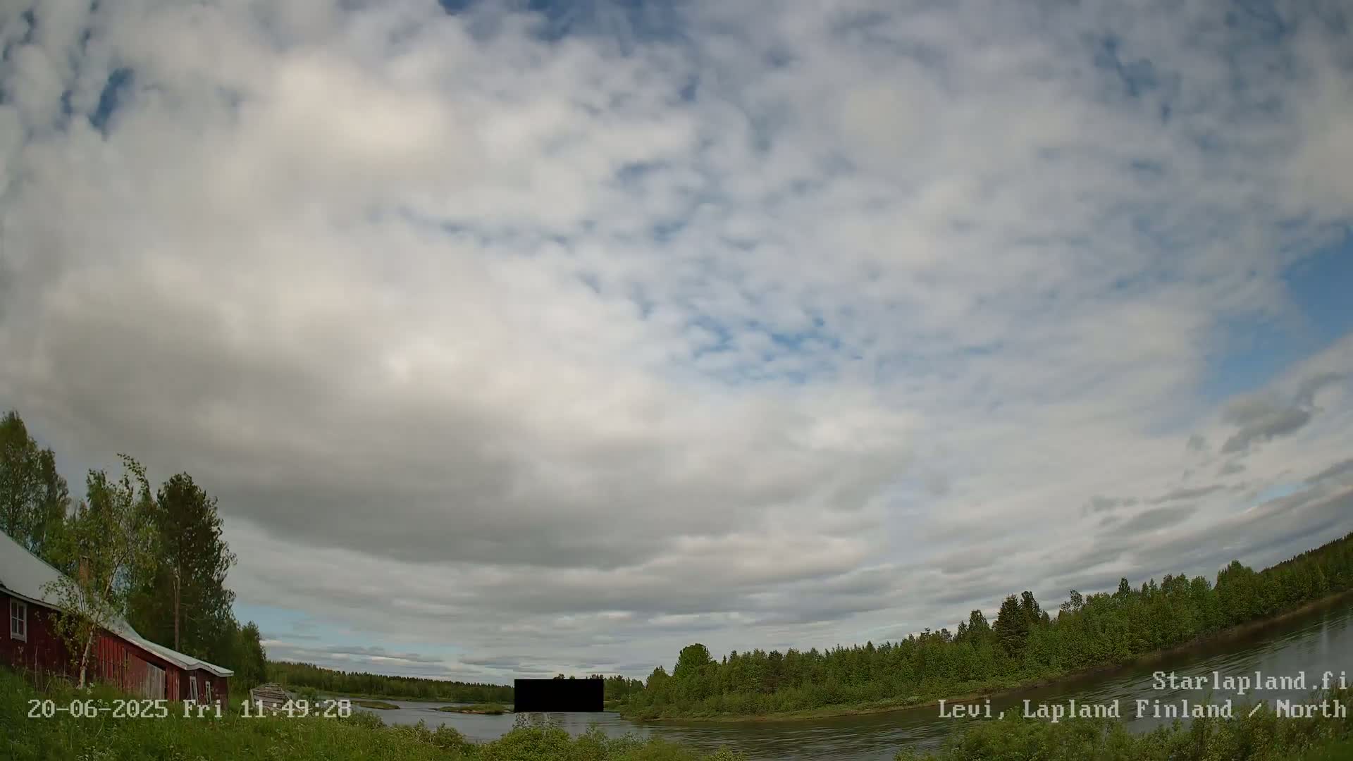 A partly cloudy sky overlooks a calm river bordered by green trees and a red building.
