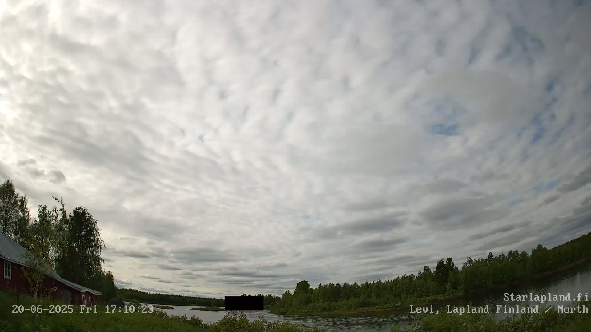 A mostly cloudy sky hangs over a tranquil river scene with a red building and green trees lining the banks.