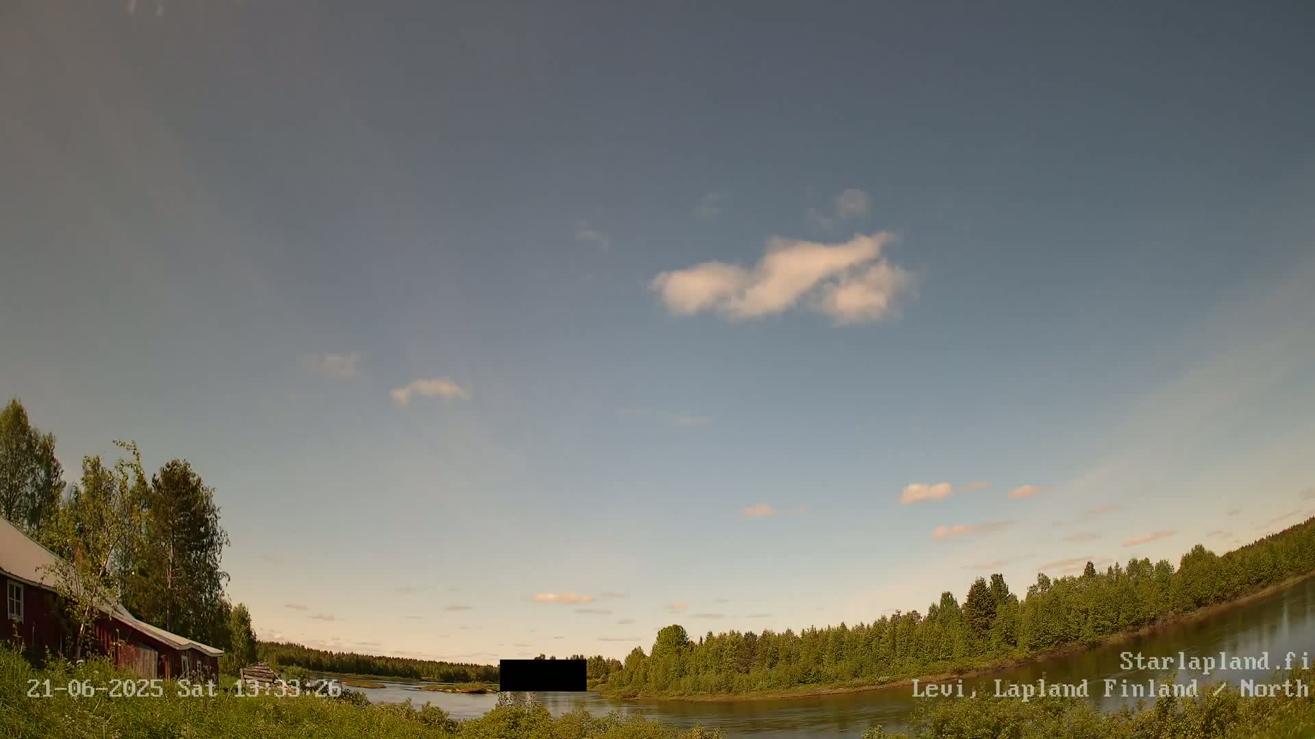 A mostly clear sky with a few clouds hangs over a calm river bordered by lush green trees and a red building on the left.