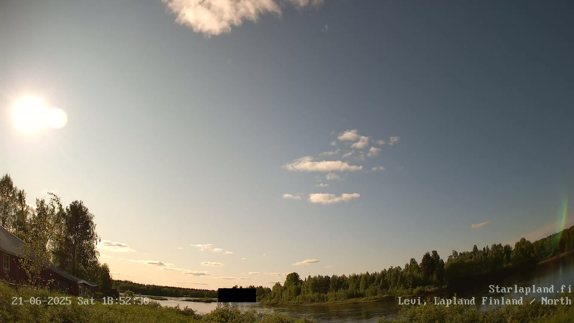 A partly sunny sky overlooks a calm river bordered by green trees and a small building near the water's edge.