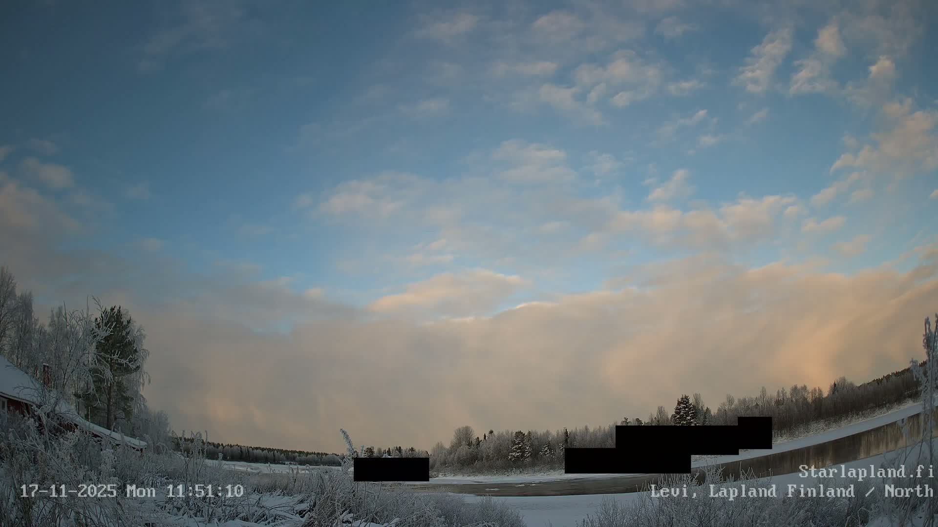 A tranquil winter landscape features snow-covered trees and ground flanking a partially frozen river, all beneath a partly cloudy sky with blue patches and clouds illuminated by the soft, golden light of a low sun.