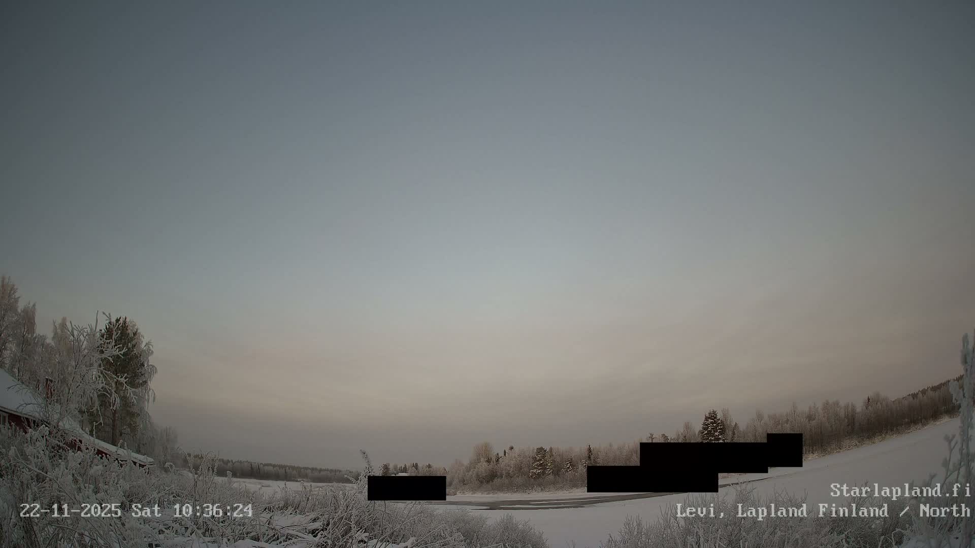A wide, snow-covered winter landscape under a muted, overcast sky reveals frosted trees, a partially frozen river, and a glimpse of a red building to the left, indicating cold and frosty conditions.