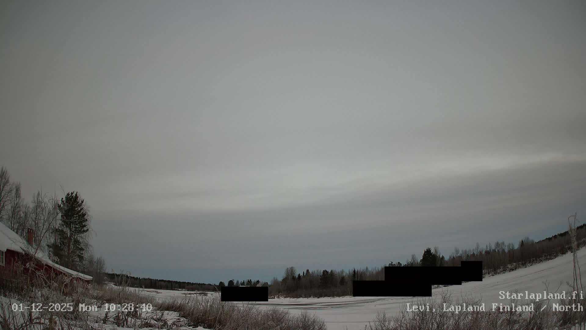 A wide-angle view captures a snow-covered winter landscape featuring a red building and trees on the left, a frozen expanse in the middle, and distant dark forests under an overcast grey sky.