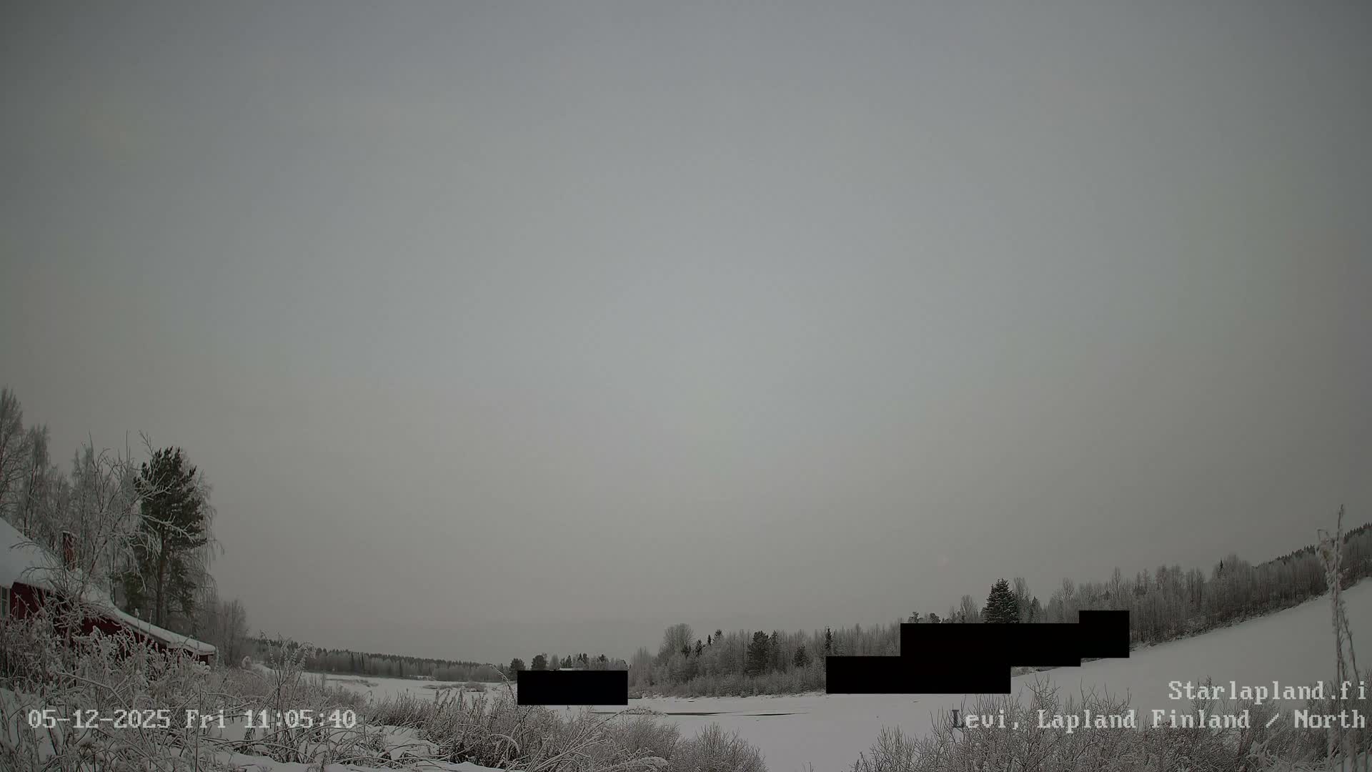A wide, wintry landscape features snow-covered ground, frosted trees, a partially frozen body of water, and a red cabin on the left under an overcast, grey sky.