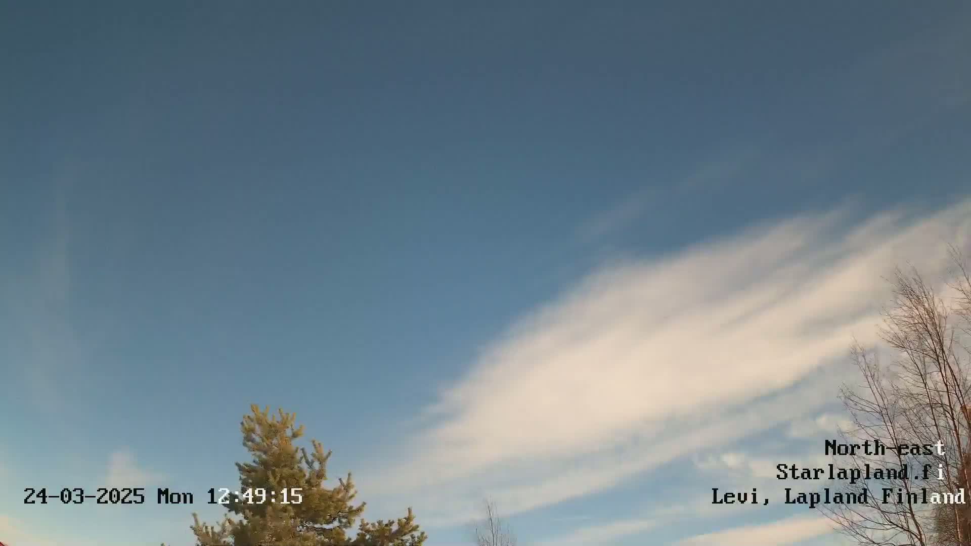 A mostly clear blue sky with thin, wispy clouds and some bare trees visible at the bottom of the frame.