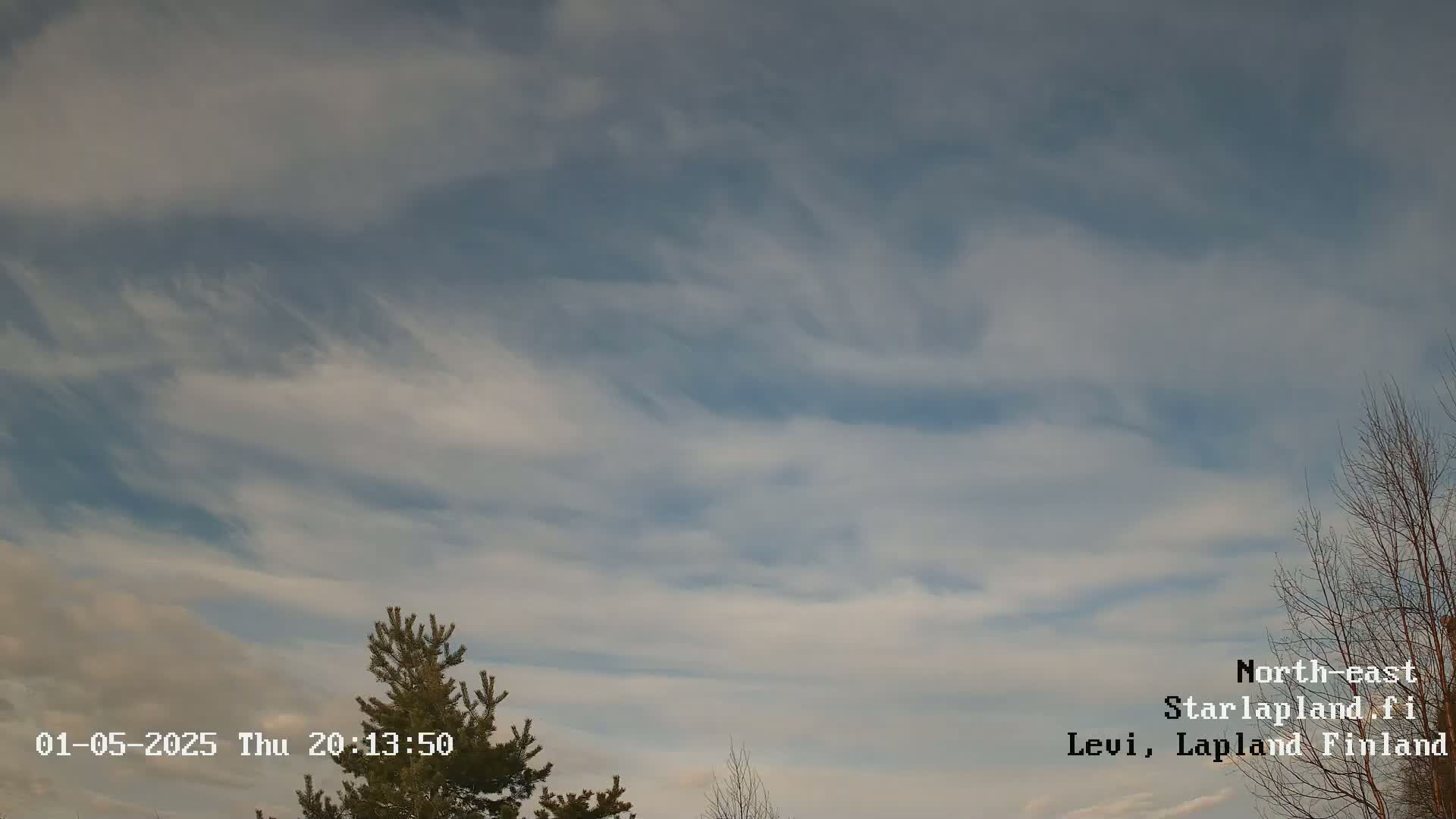 A mostly cloudy sky with wispy, high-altitude clouds and a few bare trees visible at the bottom of the frame.