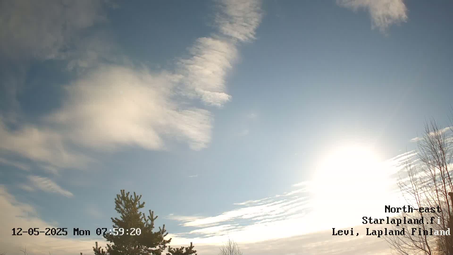 A mostly sunny sky with wispy clouds and a few bare trees is visible at the bottom of the frame.