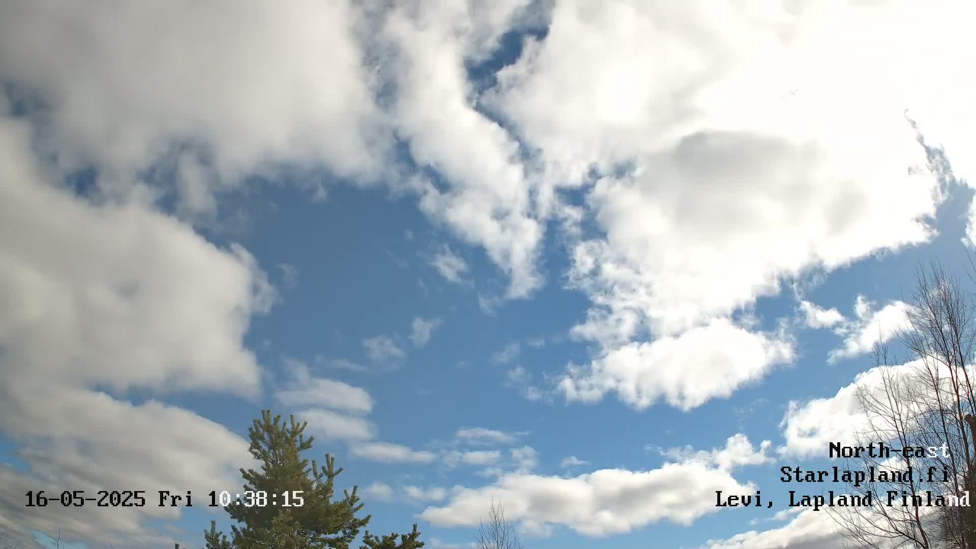 A partly cloudy sky with fluffy white clouds scattered across a mostly blue sky, partially obscuring the view of some bare trees.